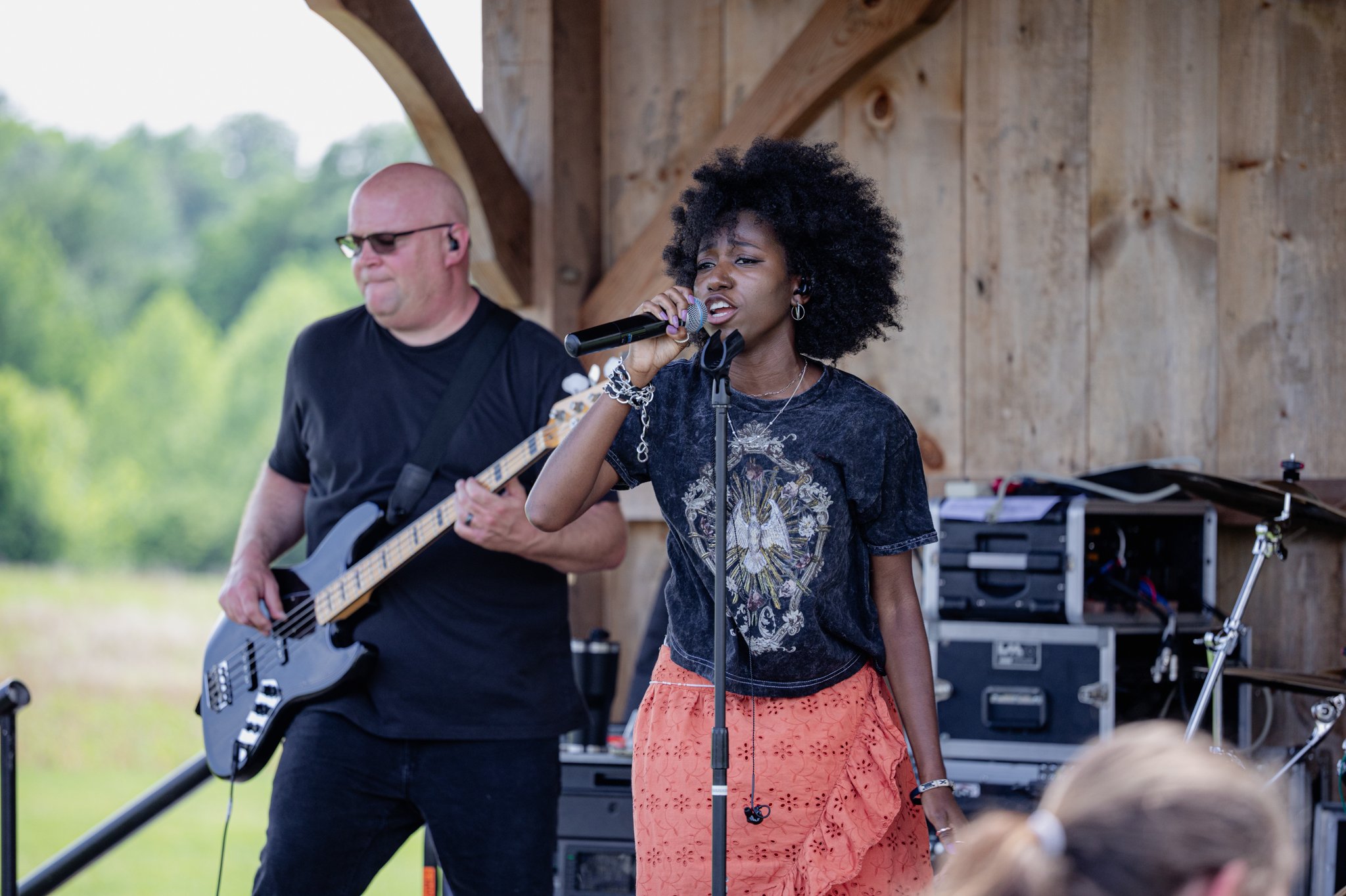 A woman with curly black hair singing into a microphone while a man is playing an electric guitar adjacent to her during an outdoor performance at a rustic wooden stage.