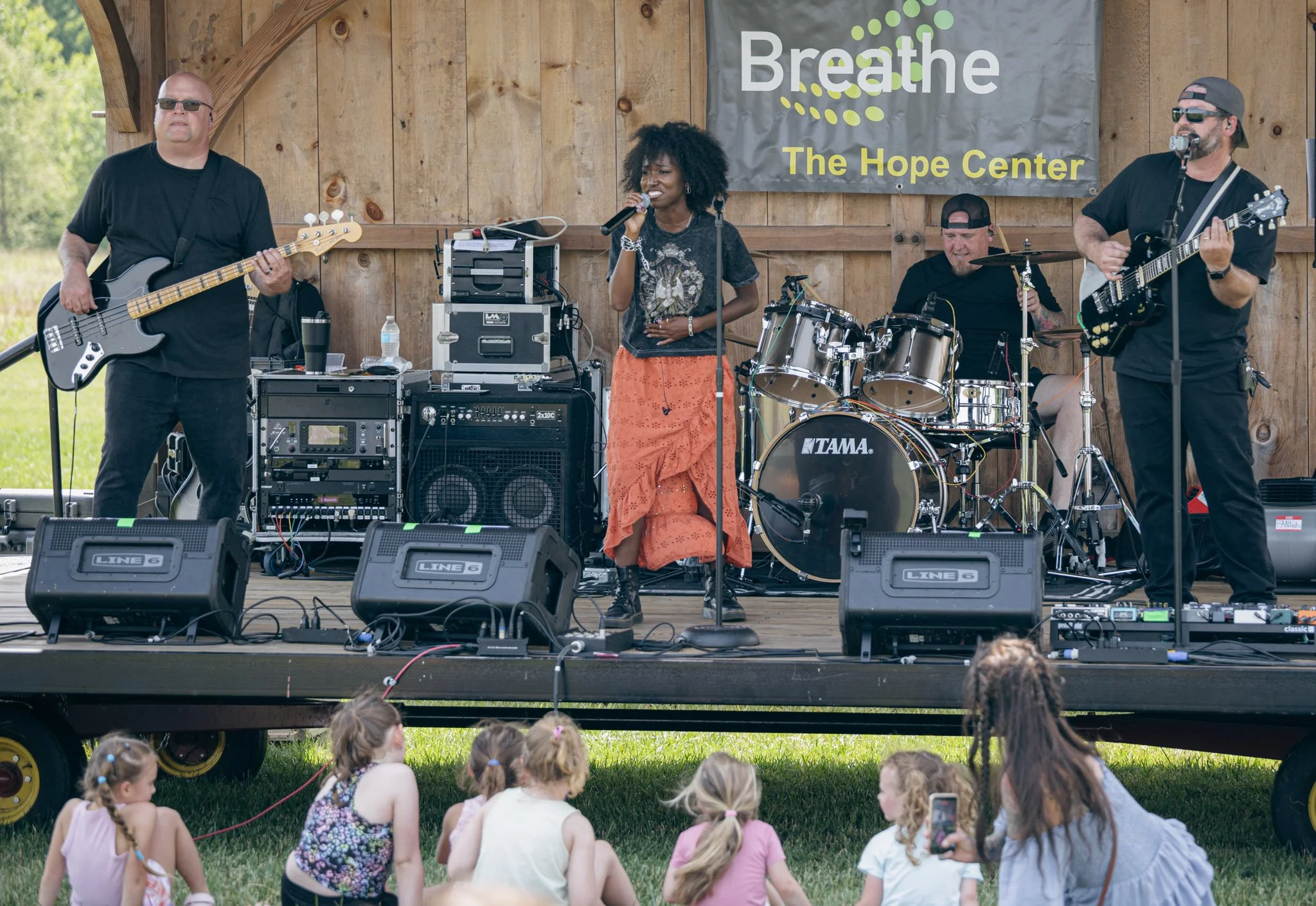 A live outdoor musical performance on a stage with four musicians and a banner that says 'Breathe The Hope Center'. The audience includes young children sitting on the grass, watching and listening to the band.