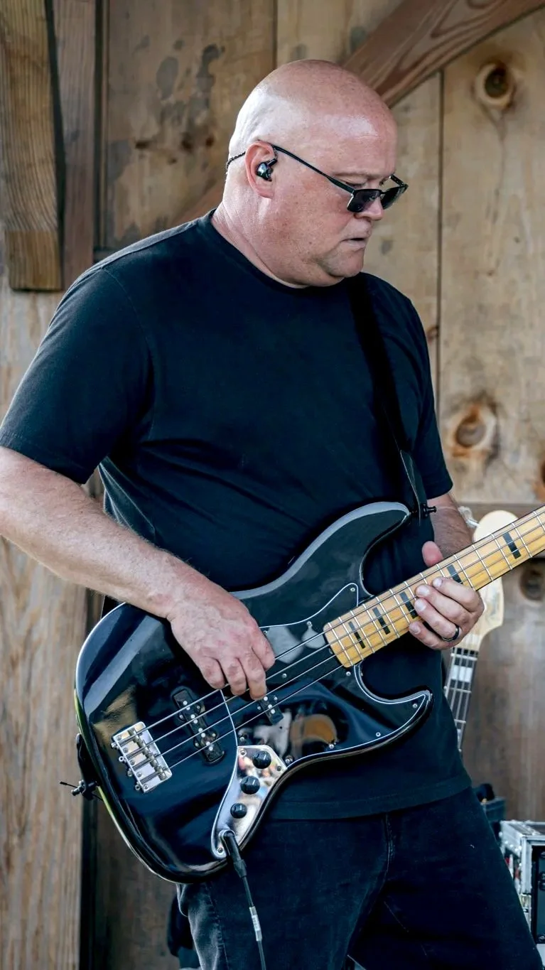 A bald man wearing sunglasses and a black T-shirt playing a black electric bass guitar with a light-colored fretboard, in a wooden setting.