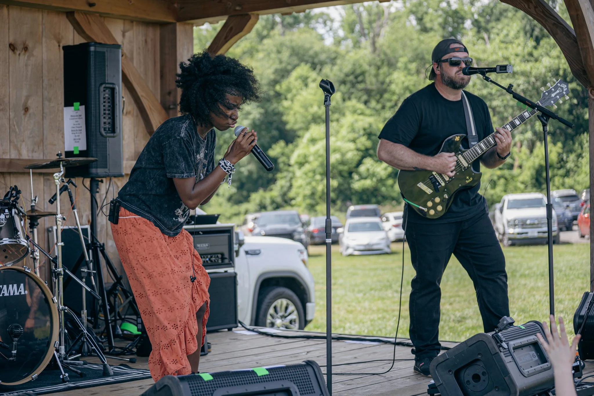 Two musicians performing on an outdoor wooden stage with musical instruments and equipment, including a drum set and a speaker, with a backdrop of green trees and parked cars.