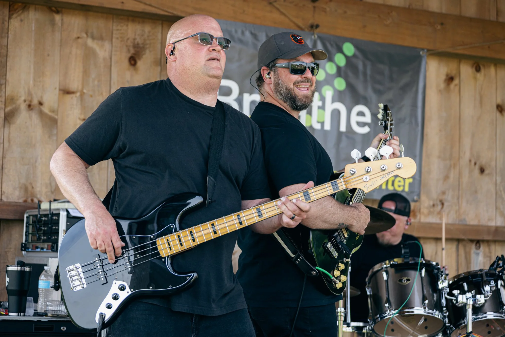 Two men performing music on stage at an outdoor event. The man on the left is playing an electric bass guitar, wearing sunglasses, a black shirt, and has a bald head. The man on the right is playing an electric guitar, wearing sunglasses, a gray cap,