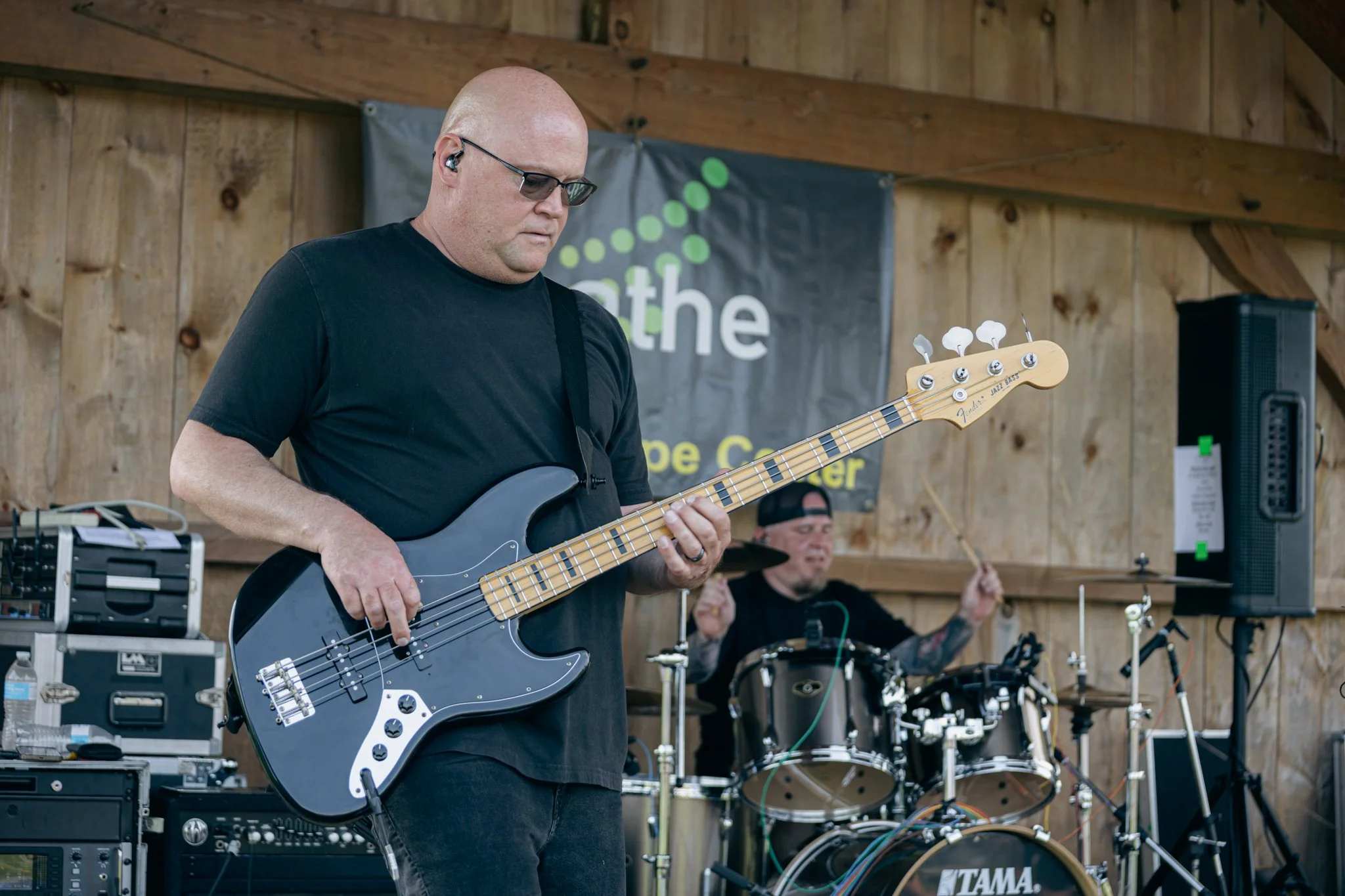 A man wearing sunglasses playing a black electric bass guitar during a live outdoor music performance, with a drummer in the background, wooden stage setting and sound equipment around.