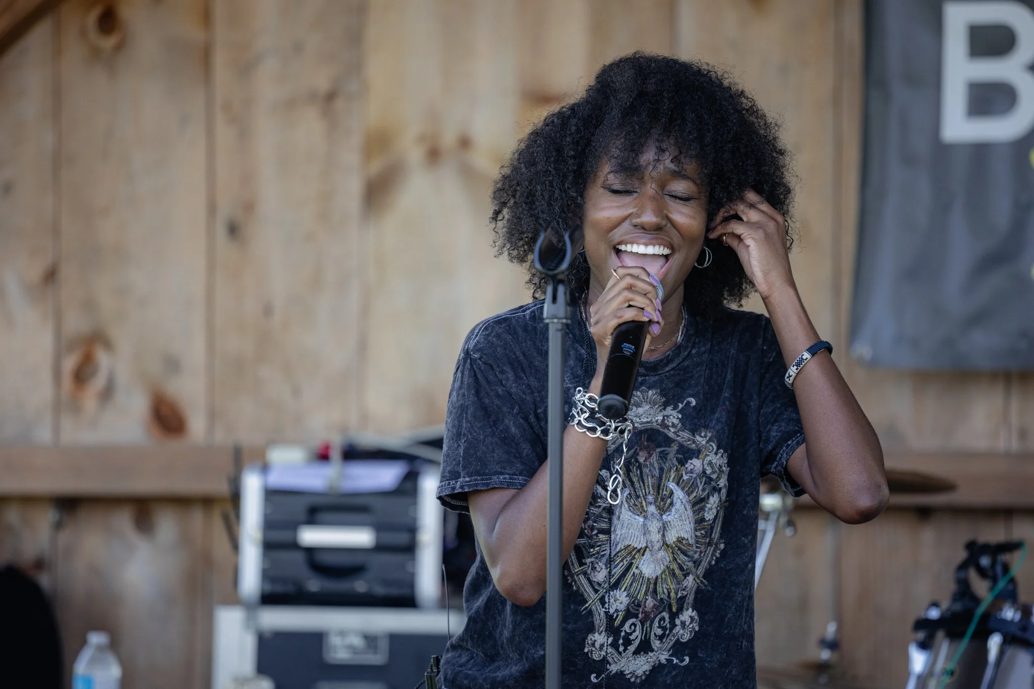 A woman with curly black hair and hoop earrings singing into a microphone indoors with a wooden wall background.