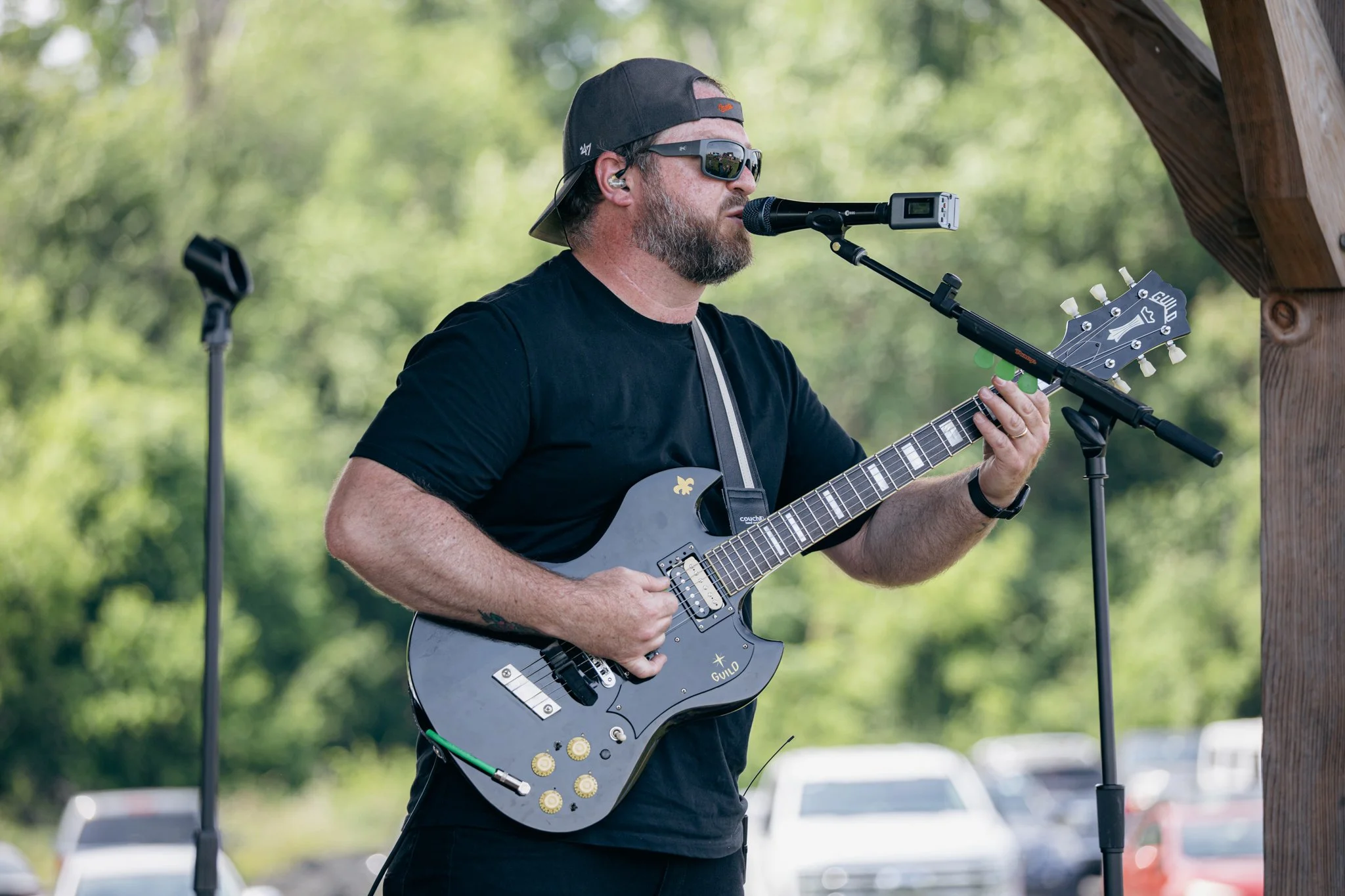 A man with a beard wearing sunglasses and a backwards black cap playing an electric guitar at an outdoor event.