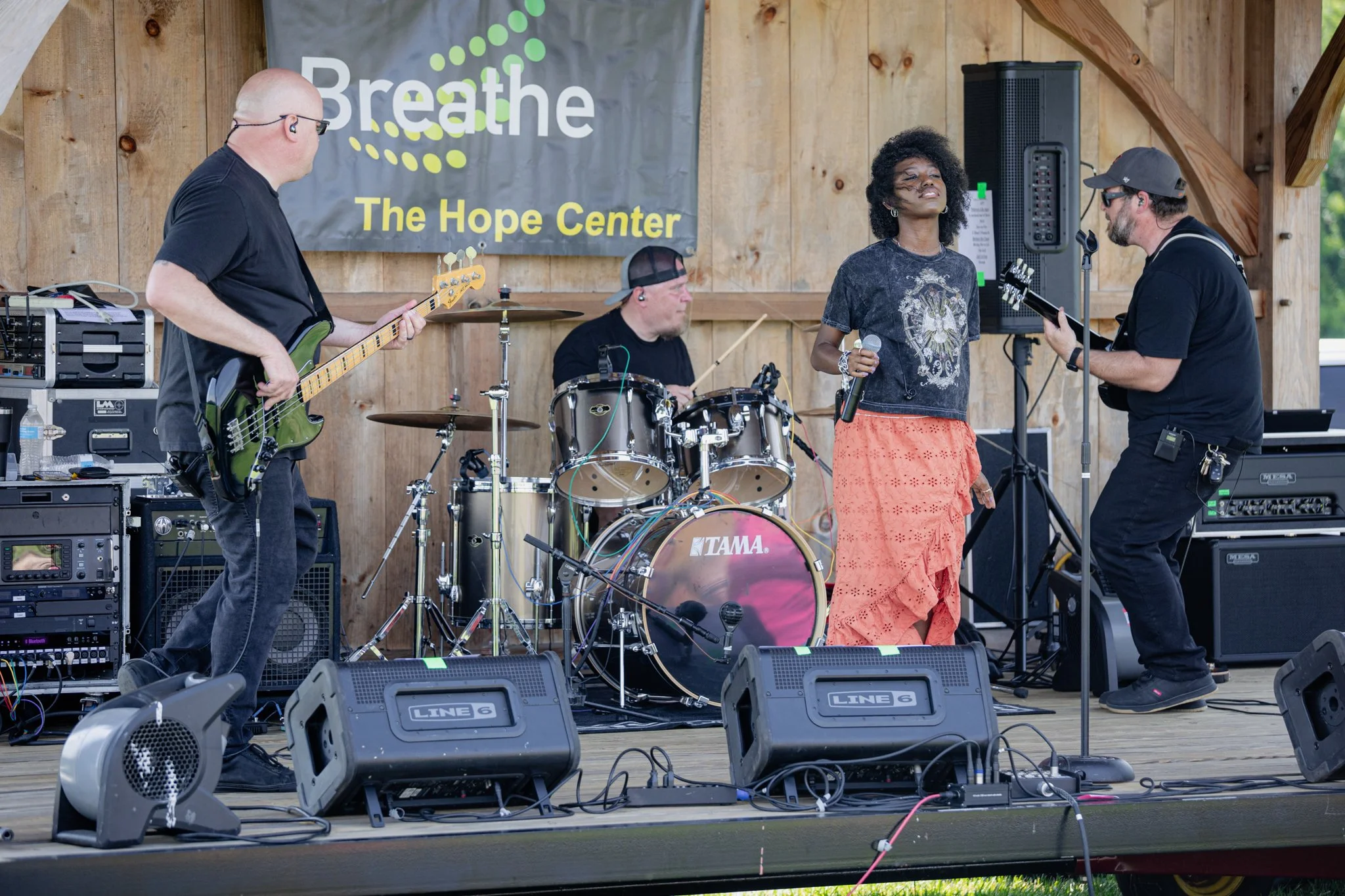 A musical performance on an outdoor stage with a band of four musicians. One woman is singing into a microphone and wearing a black graphic T-shirt and orange skirt. Three men are playing instruments: one on bass guitar, one on drums, and one on an e