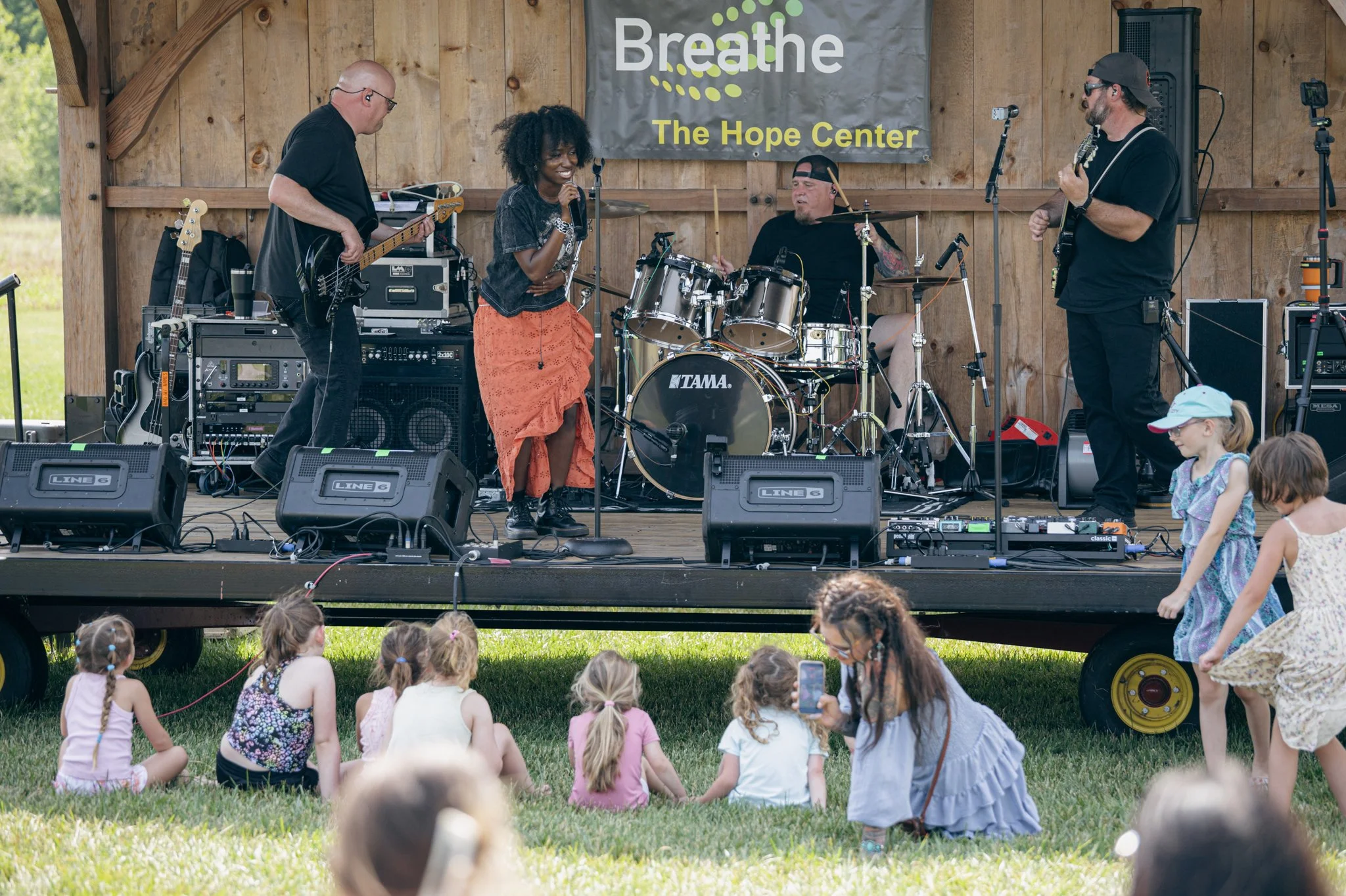 A band performs on an outdoor stage with a wooden backdrop and a banner that reads 'Breathe The Hope Center.' The band includes a woman singing into a microphone, a man playing electric guitar, a man playing drums, and another man playing a guitar. C