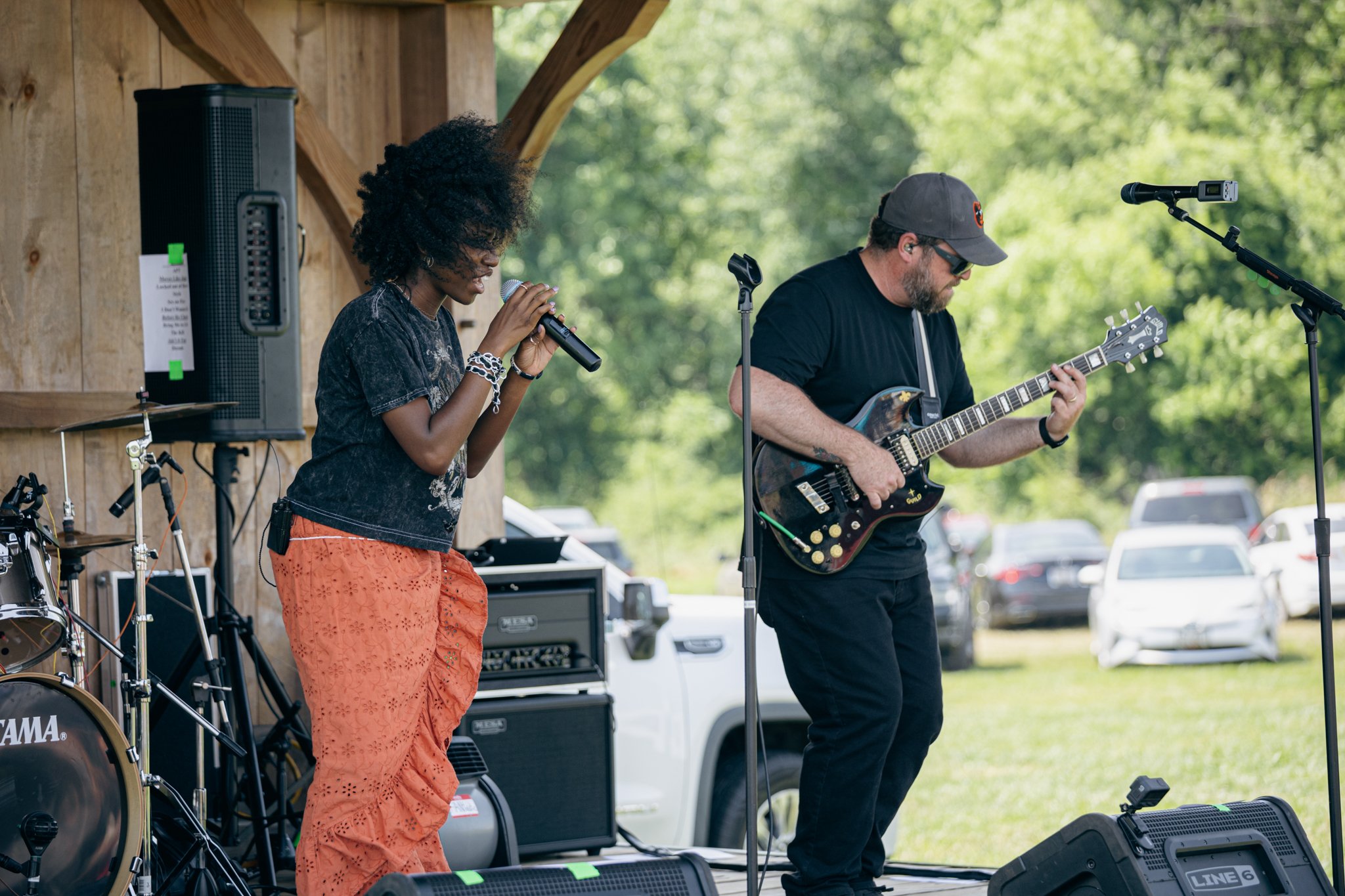 Two musicians performing outdoors under a pavilion. One woman with curly hair, wearing an orange skirt and a black top, singing into a microphone. One man with a beard, wearing a gray cap, sunglasses, black shirt, playing an electric guitar.
