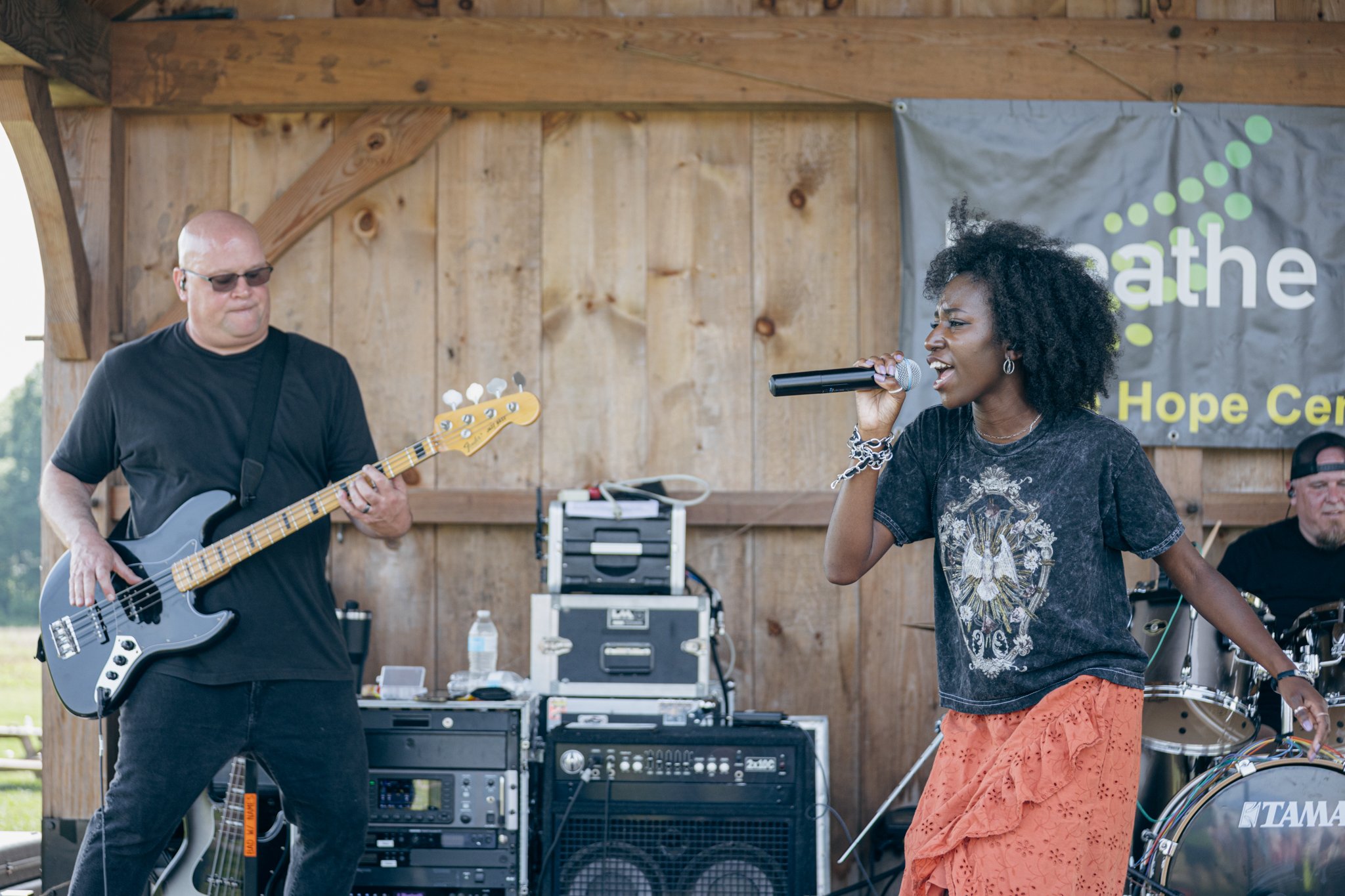 A woman singing into a microphone and a man playing a bass guitar performing on an outdoor stage with a wooden backdrop and sound equipment.