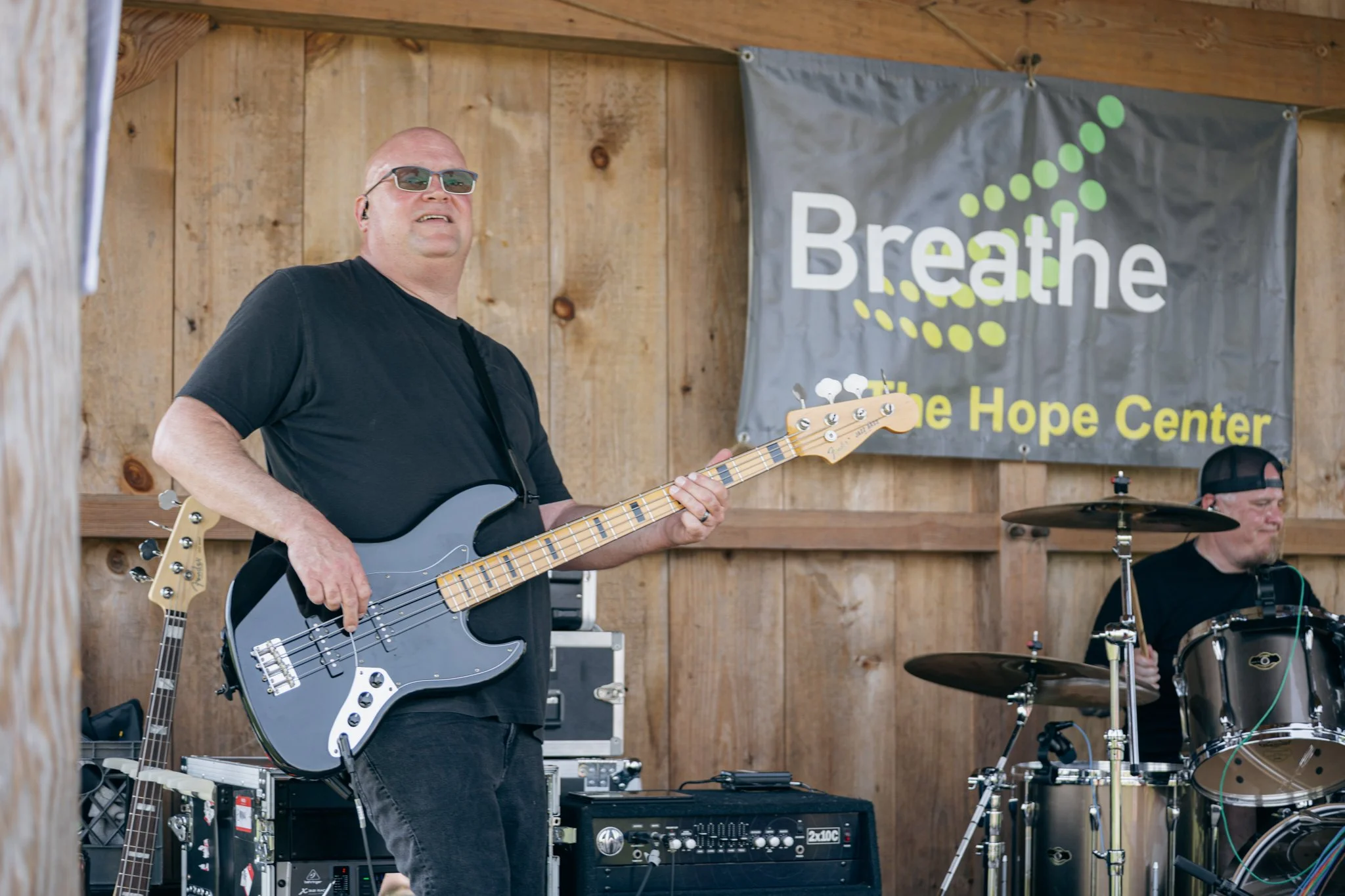 A man wearing sunglasses playing an electric guitar on a wooden stage at an outdoor event, with a drum set and other musical equipment visible in the background. A large banner behind him reads 'Breathe The Hope Center'.