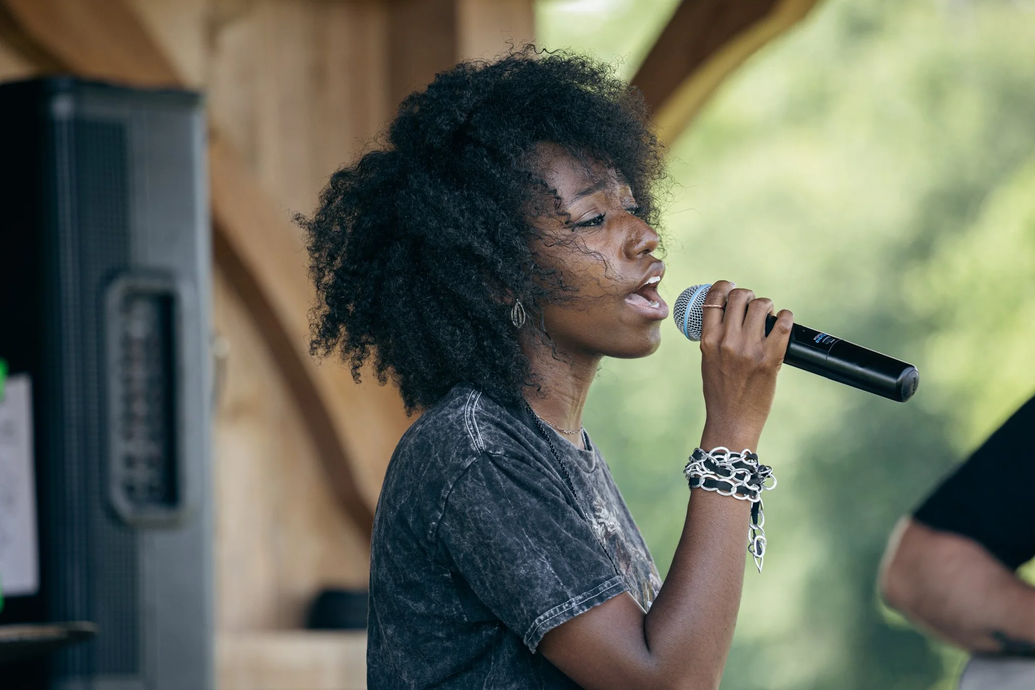 Young woman with curly black hair singing into a microphone outdoors.