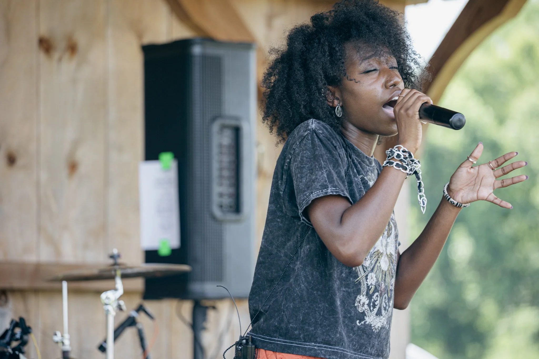 A woman singing into a microphone with her eyes closed, wearing earrings, bracelets, and a dark t-shirt, in an outdoor setting with wooden walls and a speaker in the background.