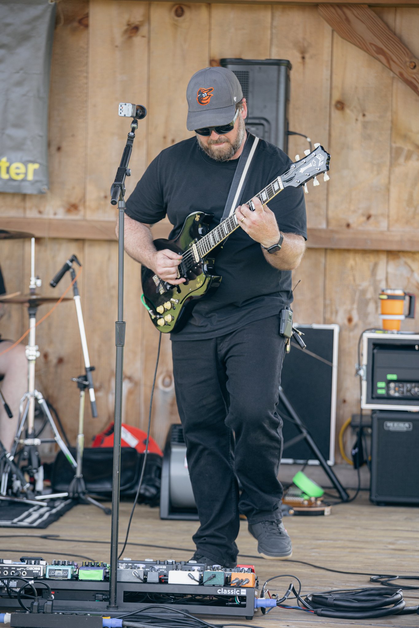 A man wearing sunglasses, a gray cap with a Baltimore Orioles logo, black t-shirt, and black pants playing an electric guitar on a wooden stage. There are musical equipment and amplifiers visible around him.