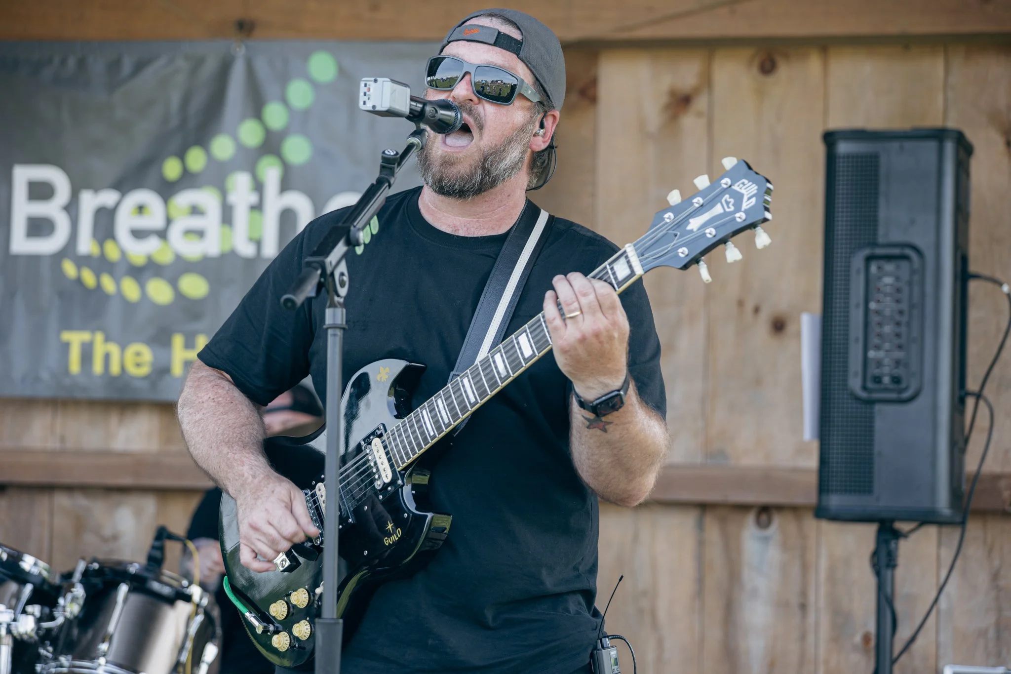 Man with gray beard wearing sunglasses and a gray cap playing an electric guitar on stage, with a microphone in front of him, during a live performance.