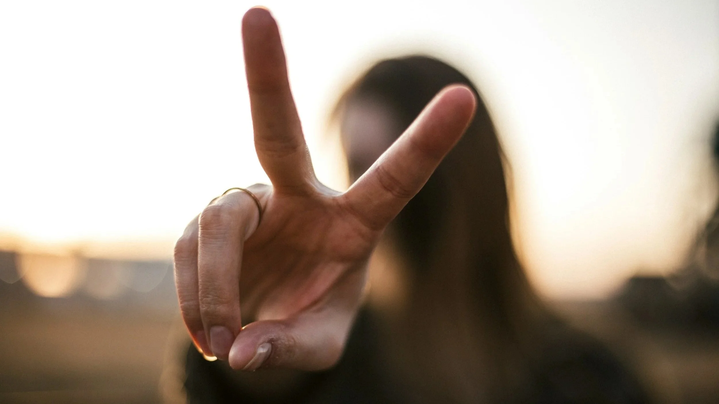 A person making a peace sign with their hand, fingers held up, in front of their face which is blurred and in shadow.