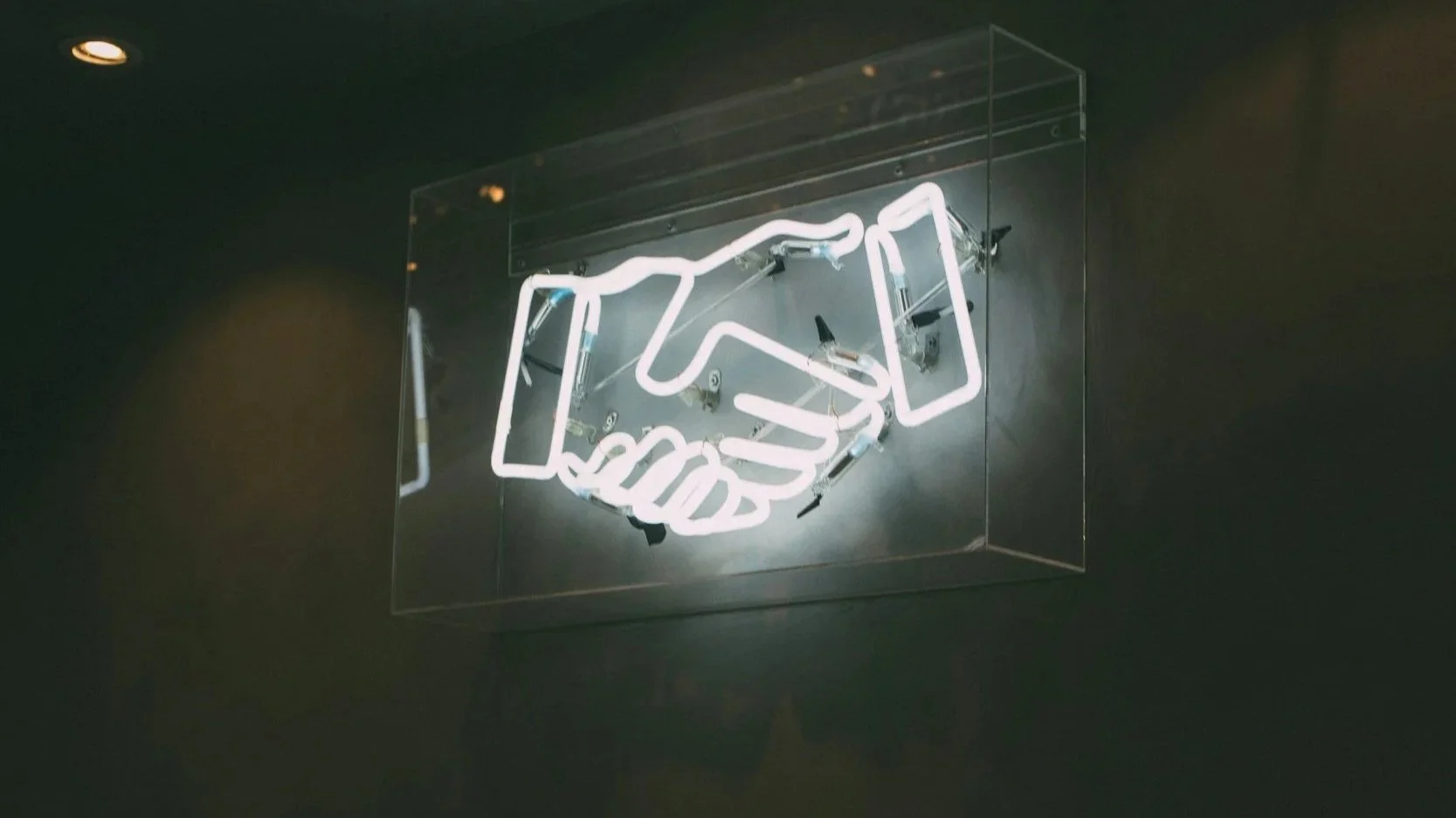 Neon sign depicting a handshake, mounted on a dark wall.