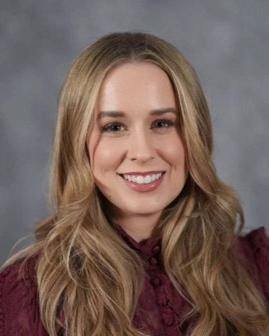 Portrait of a smiling young woman with long, wavy blonde hair wearing a burgundy top with a ruffled neckline, against a gray background.