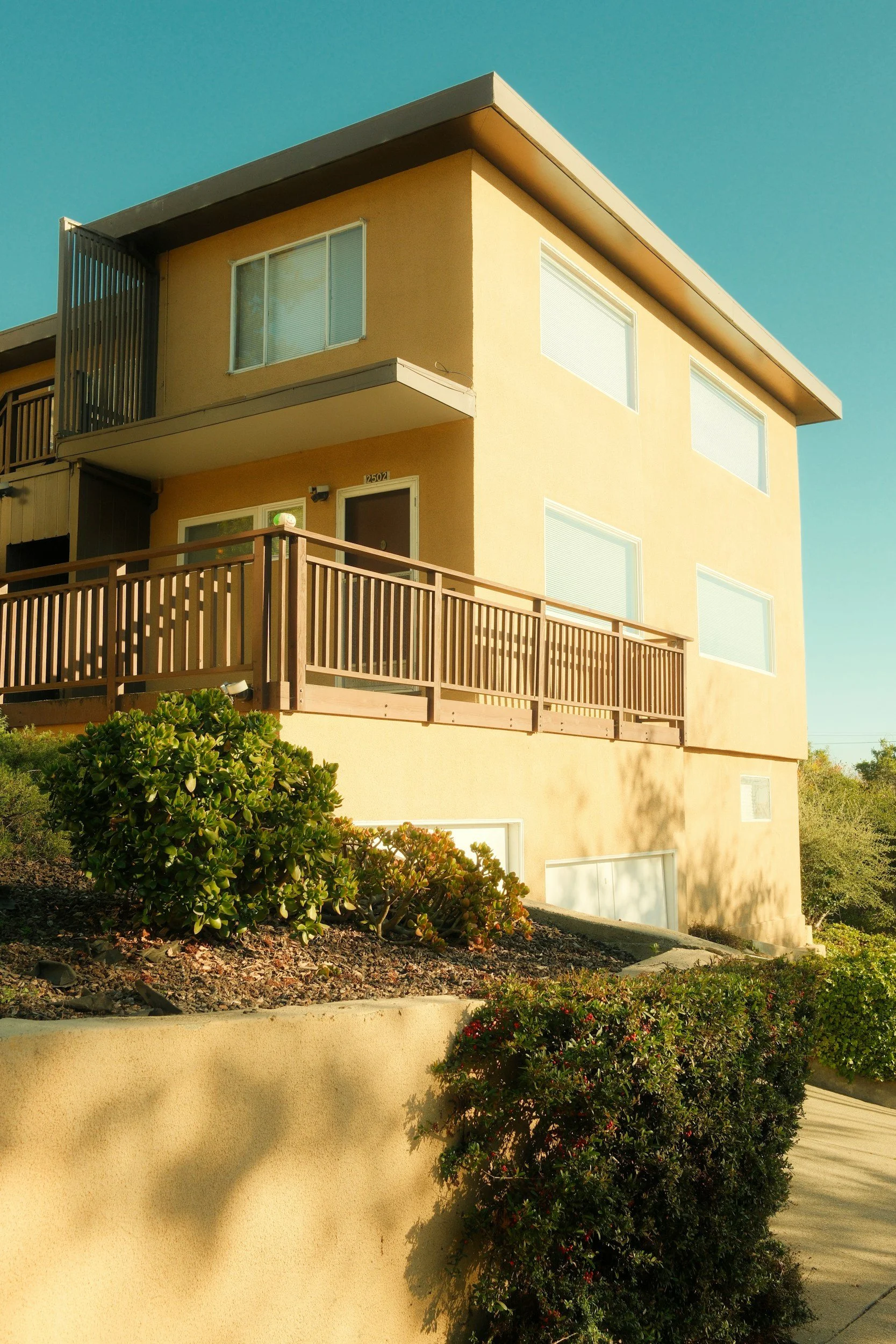 Exterior view of a three-story beige residential building with balconies, surrounded by landscaping including bushes and trees, under a clear blue sky.