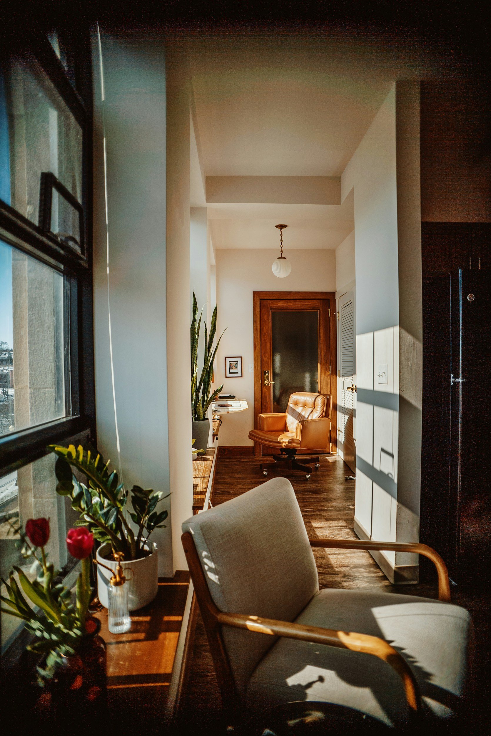 Bright sunlit corner of a cozy living space with a white chair by a wooden-edged window, potted plants, a wooden door, and a leather armchair in the background.