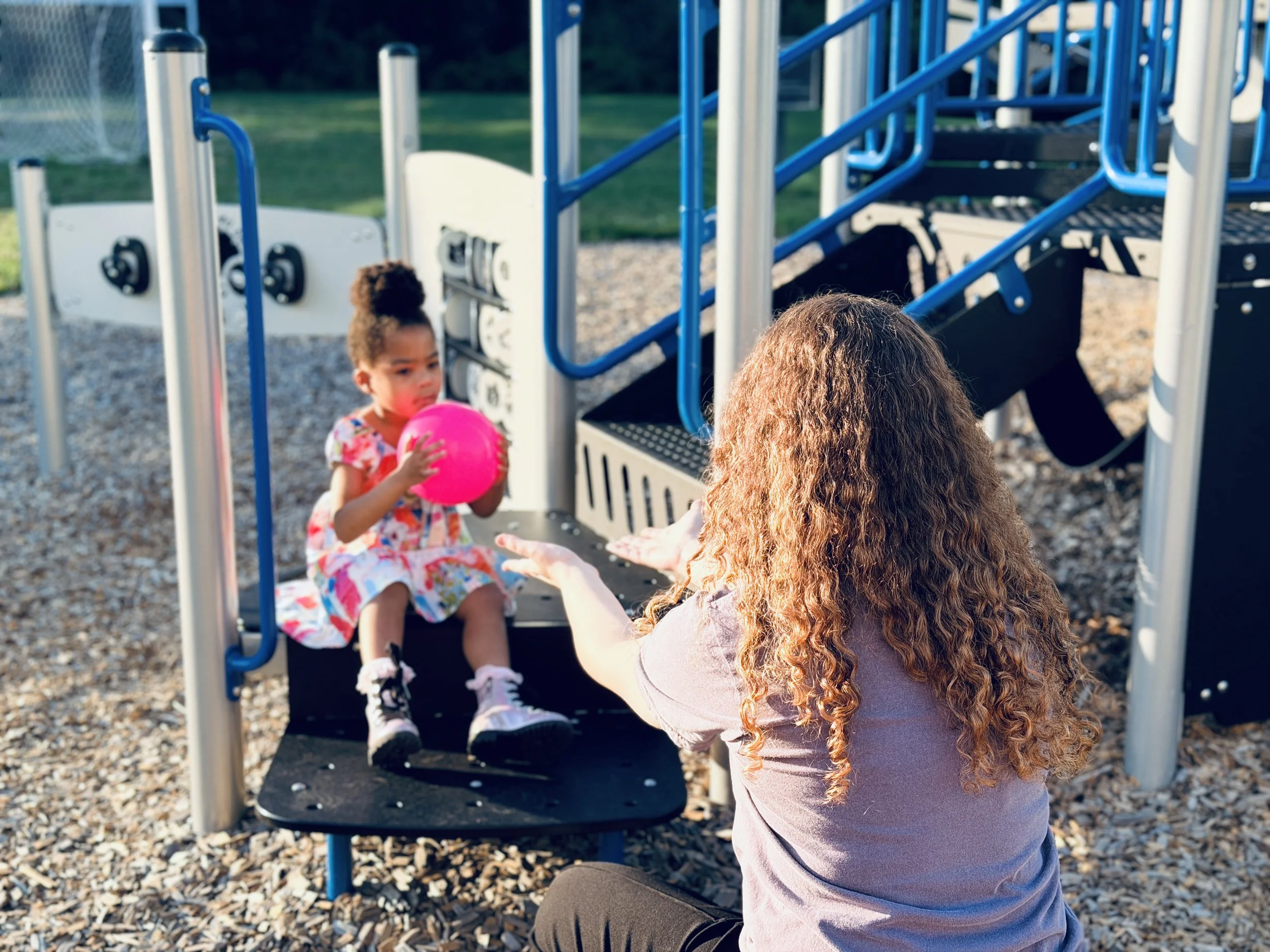 A woman with long, curly hair is reaching out to a young girl sitting on a playground structure, holding a pink ball. The girl has curly hair in a high bun and is wearing a colorful dress, with a focused expression. The playground has blue and black equipment with a gravel surface, and a grassy field in the background.