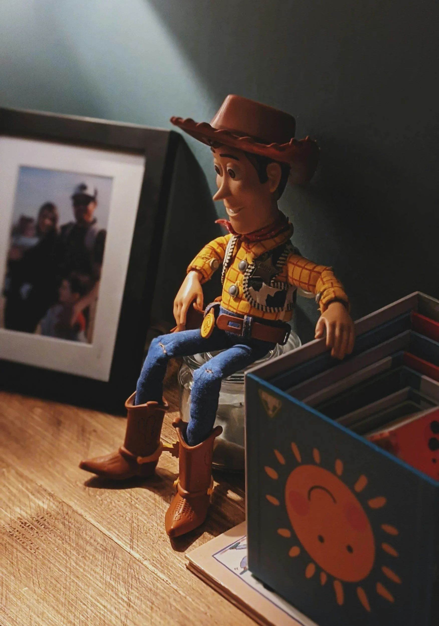 A woody doll dressed as Woody from Toy Story, wearing a cowboy hat, yellow plaid shirt, jeans, and cowboy boots, sitting on a surface next to a framed photograph and a card with a cartoon sun character.