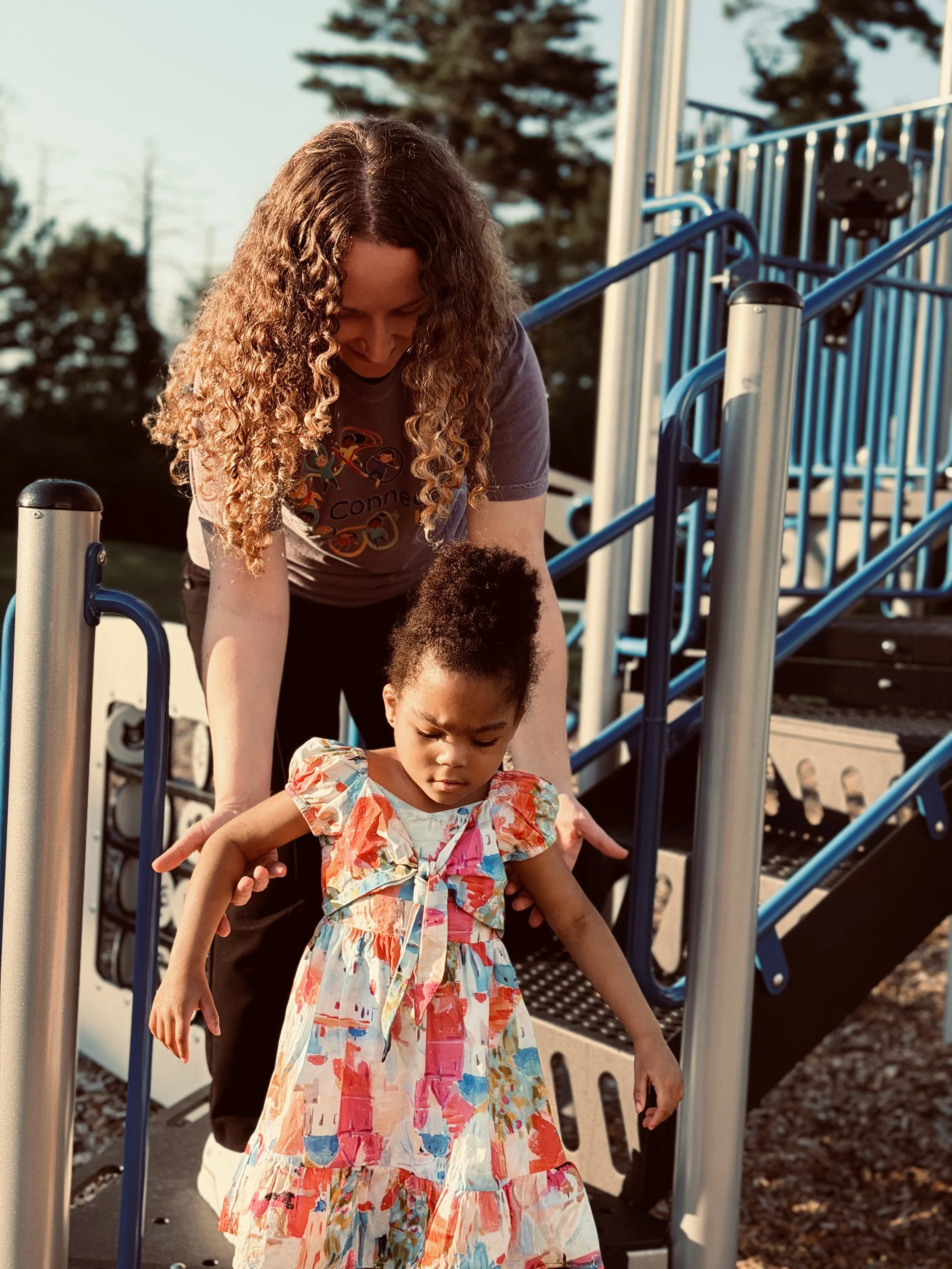 A woman and a young girl walking down playground stairs together outdoors during late afternoon or early evening, with trees in the background.