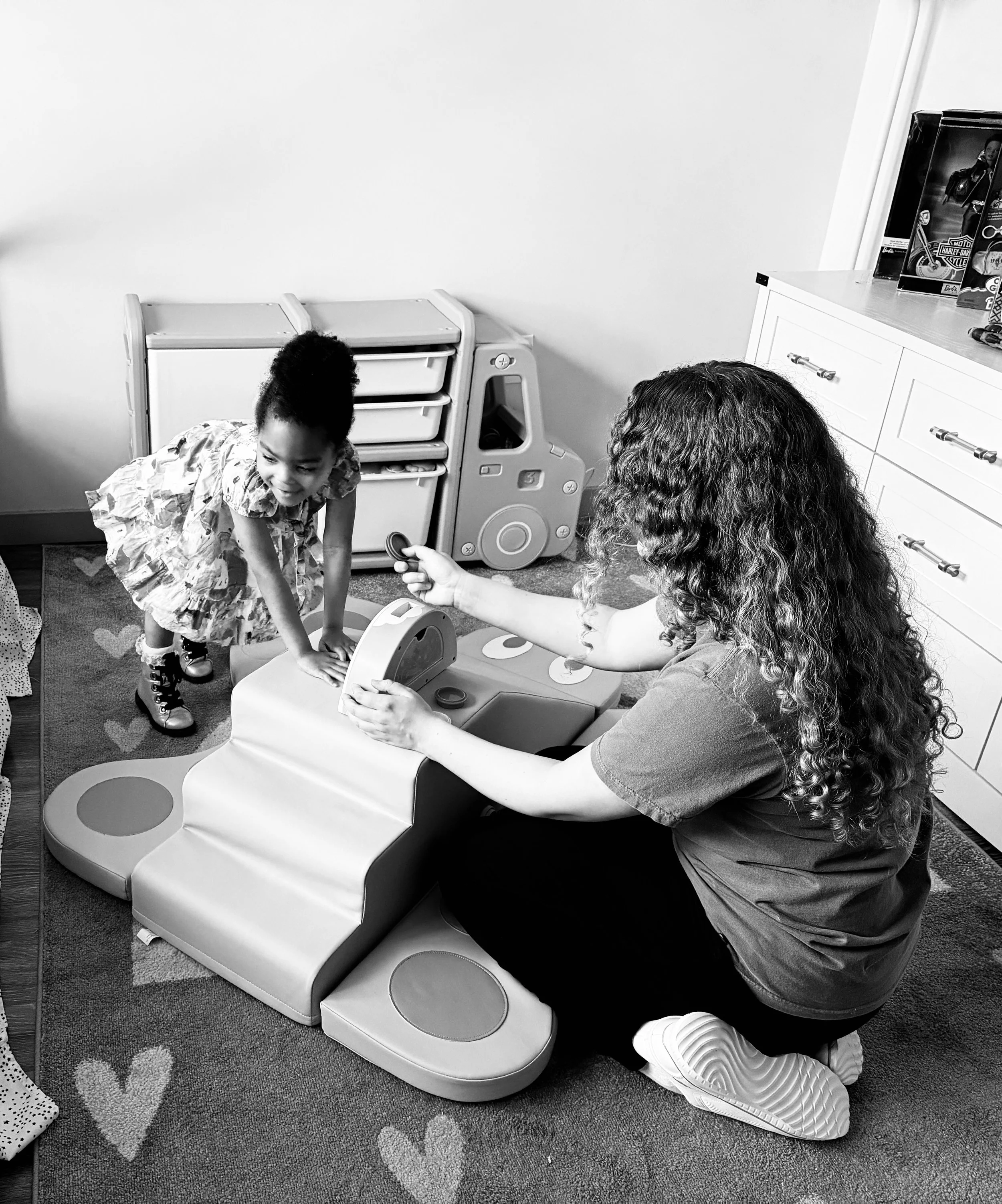A young girl in a patterned dress bending over, smiling while playing with a toy on a table, with a woman sitting next to her, holding a game piece, in a room with a cabinet and toys on top.