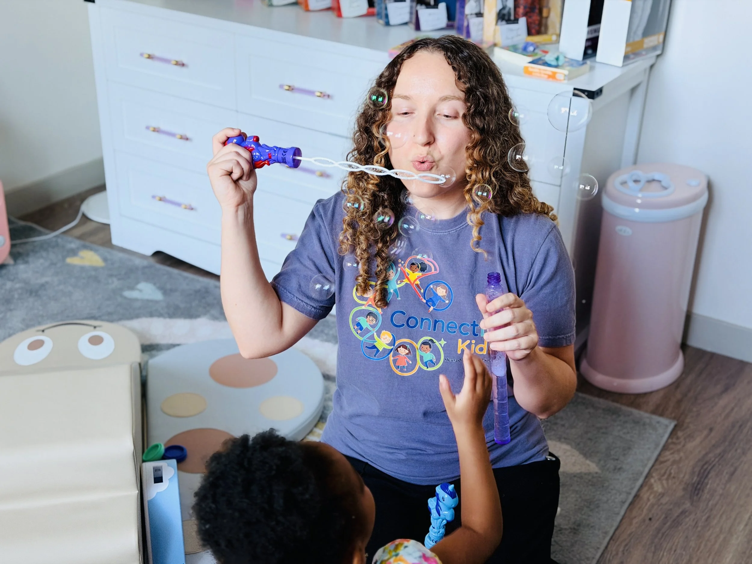 A girl with curly hair blowing bubbles in a room filled with shelves and furniture, children are visible in the foreground.