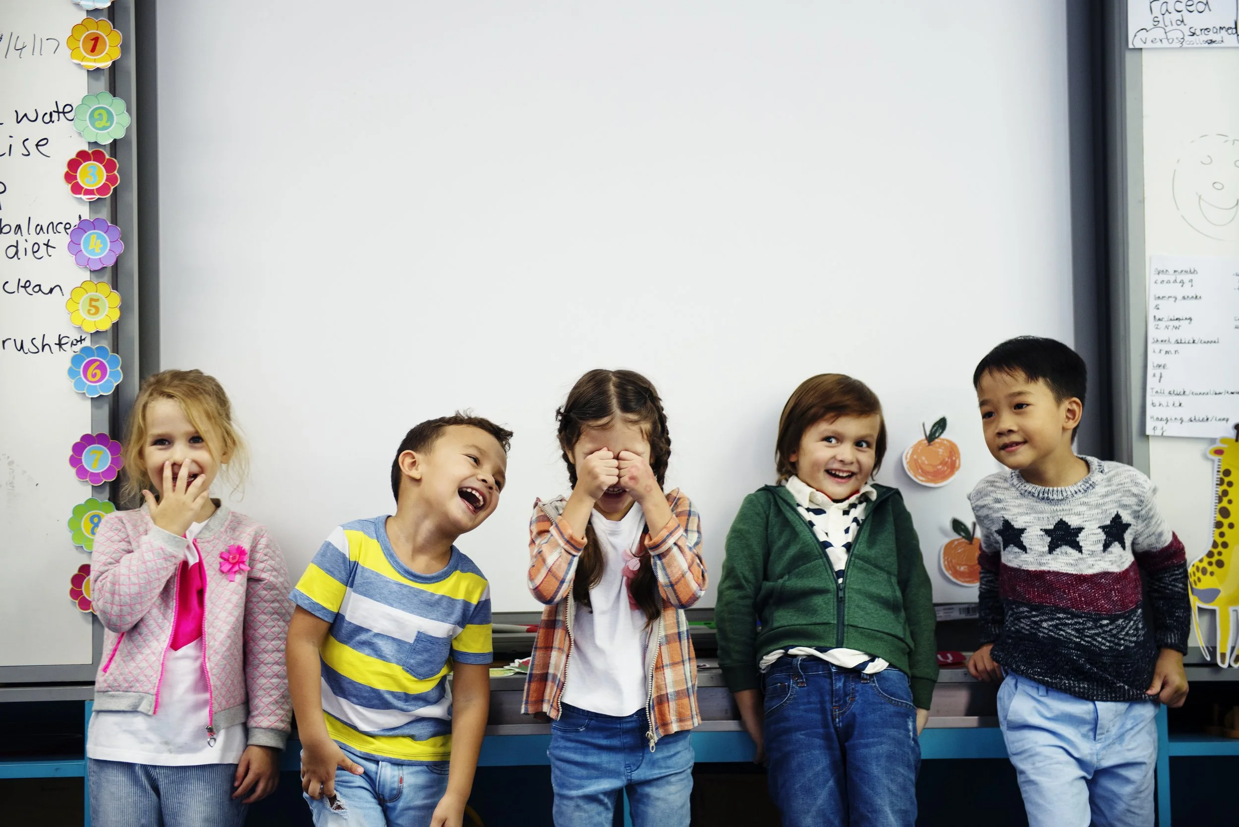 A group of six children standing in front of a classroom whiteboard, laughing and smiling.