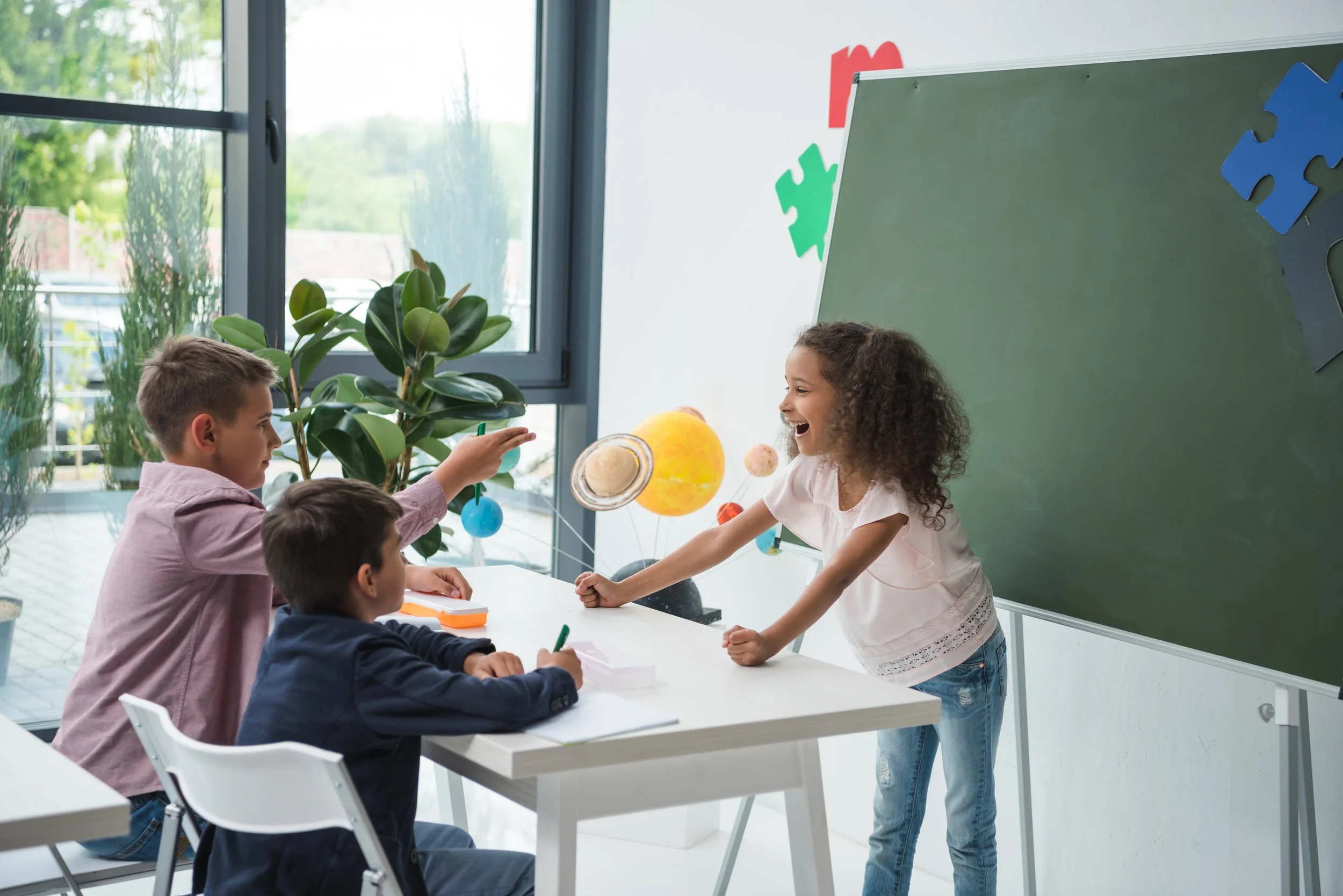 A classroom with children participating in a science activity about the solar system, with a girl gesturing towards planets and orbiting models on the table.