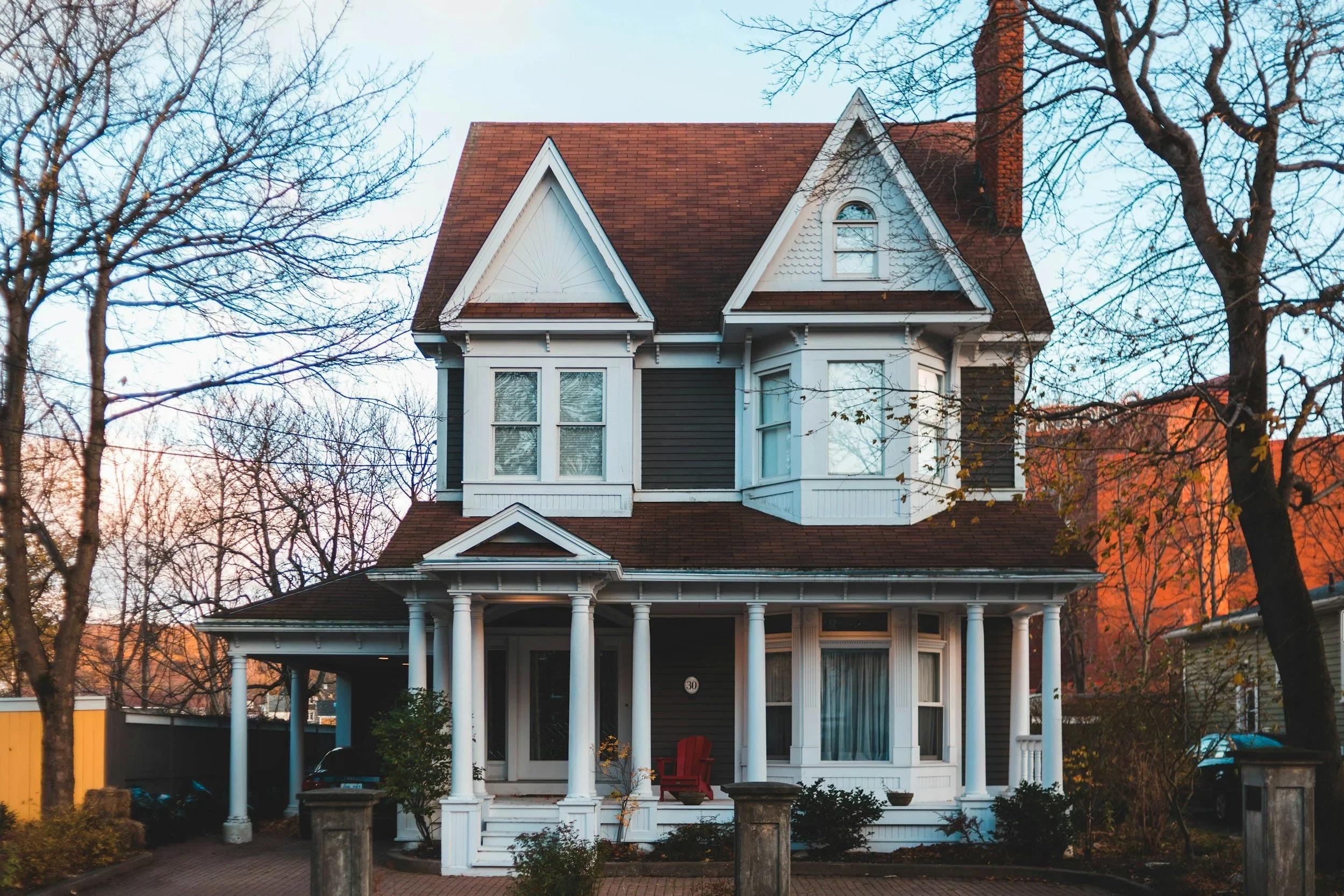 A two-story house with a front porch supported by white columns, a steep gabled roof, and bay windows, surrounded by trees with mostly bare branches during fall or winter.