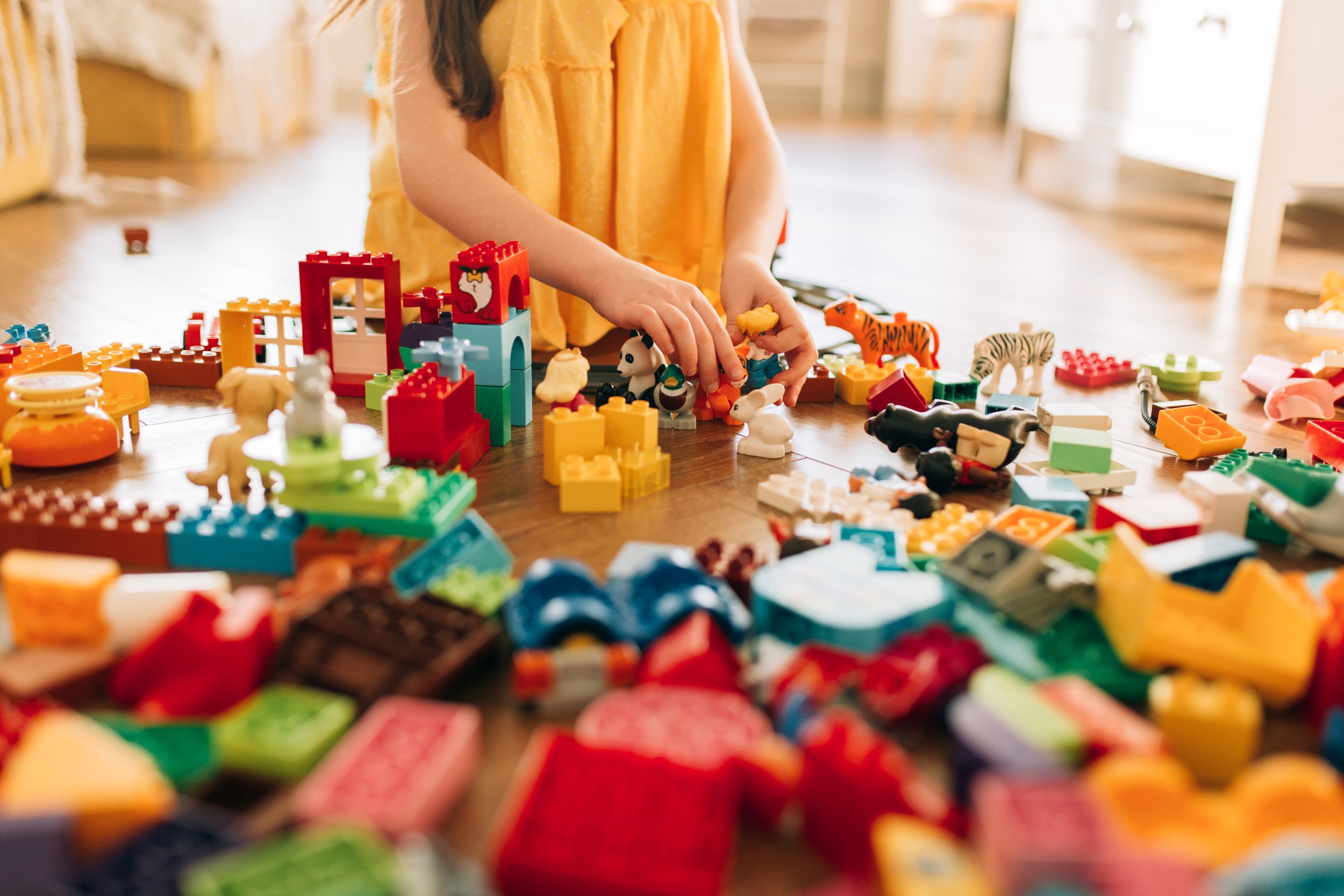 Child playing with colorful building blocks and toy animals on a wooden floor in a well-lit room.