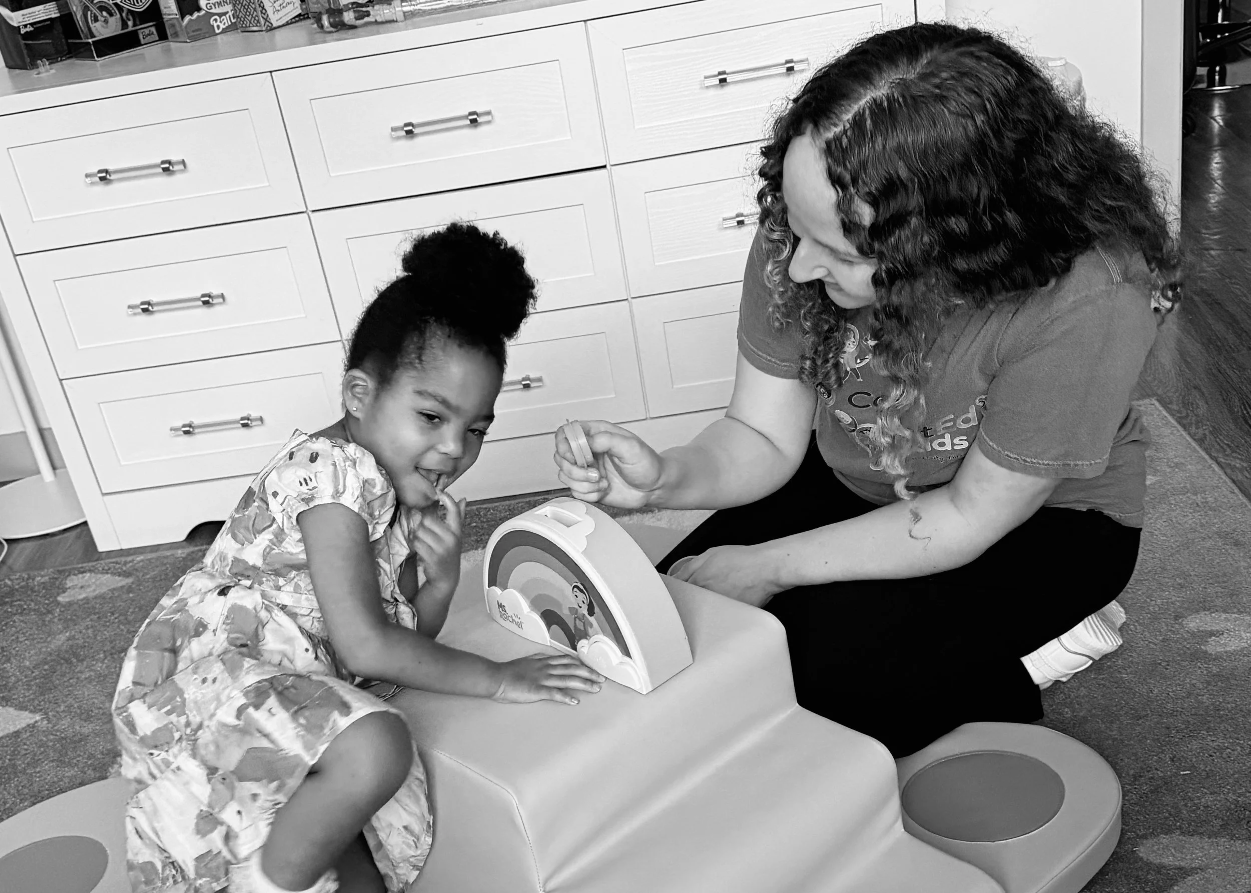 A woman and a young girl playing a game together at a table in a room with white cabinets and a bookshelf, both smiling and engaged in the activity.