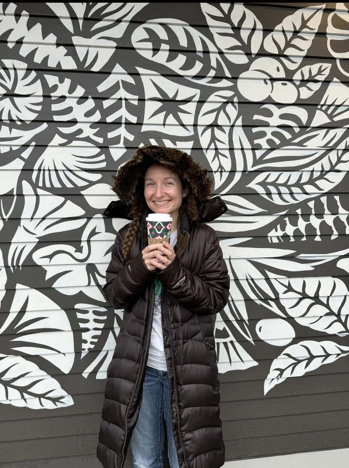 A young woman with braided hair wearing a long black puffer coat and a furry hood, holding a to-go coffee cup, standing in front of a black and white leafy mural.