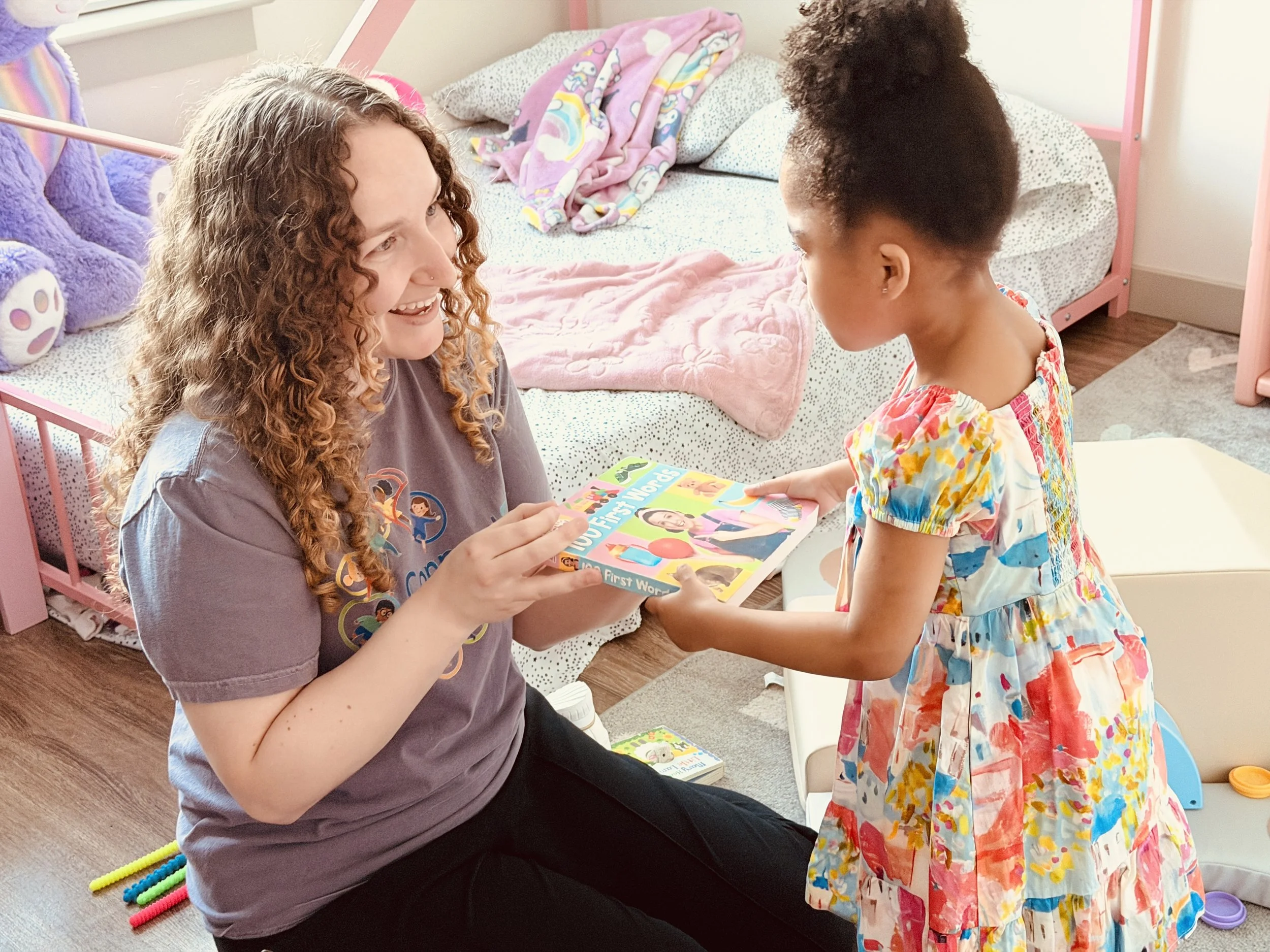 A woman with curly hair sitting on the floor holding a colorful children's book titled "First Words." A young girl with curly hair in a colorful dress is standing in front of her, both are smiling and engaging in an activity indoors, with a pink bed and blankets in the background.