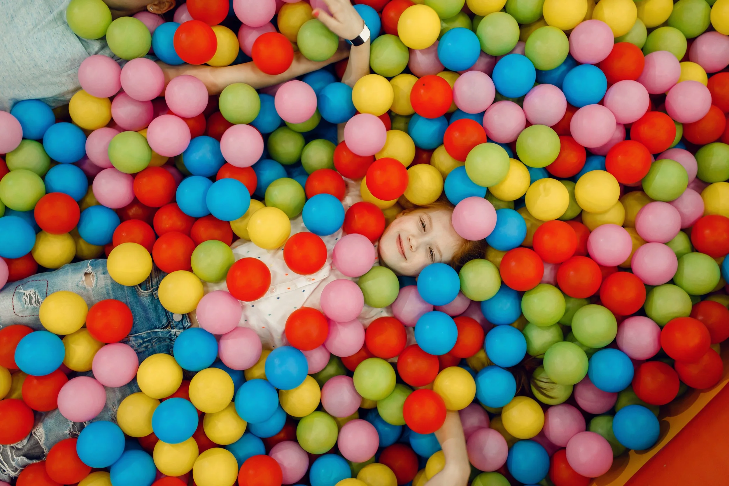 A girl lying in a colorful ball pit filled with red, blue, pink, green, and yellow balls, smiling and surrounded by the vibrant colors.