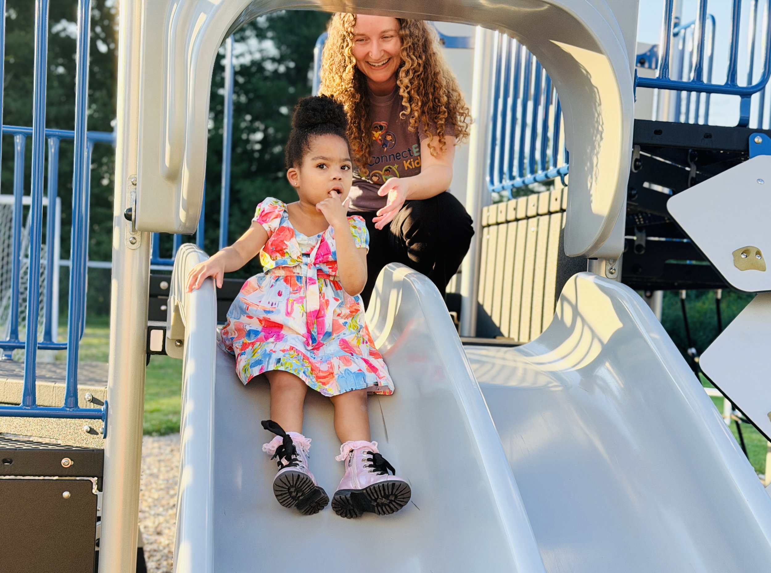 A young girl with curly hair in a colorful dress sitting on a playground slide, with a woman smiling behind her, at a park in the late afternoon.