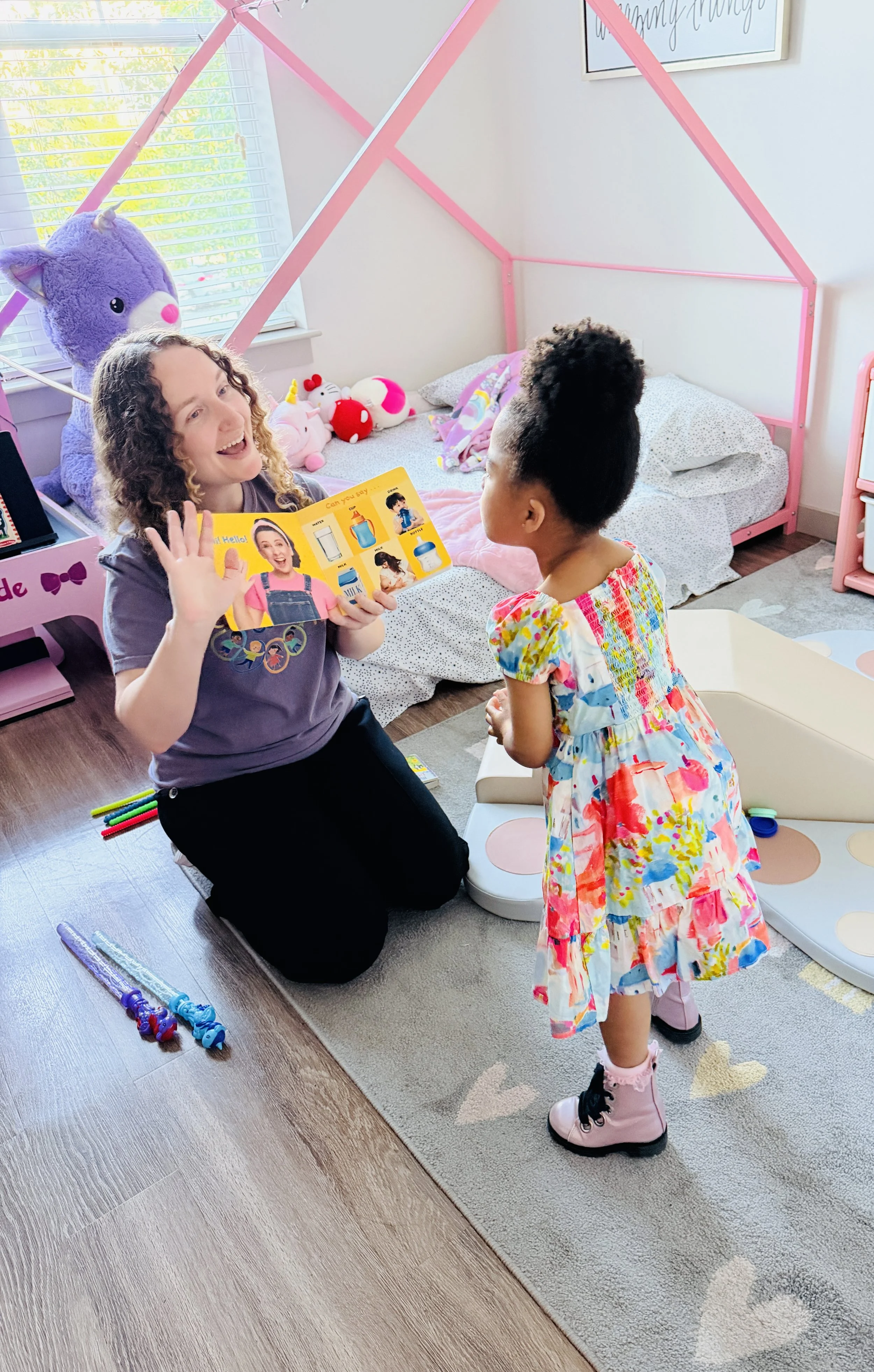 A woman kneeling on the floor and a young girl standing facing each other in a child's bedroom. The woman is smiling and holding a colorful board book. The girl is wearing a colorful dress and pink boots. The bedroom has a pink tent frame over a bed with stuffed animals, and there are toys and markers on the floor.