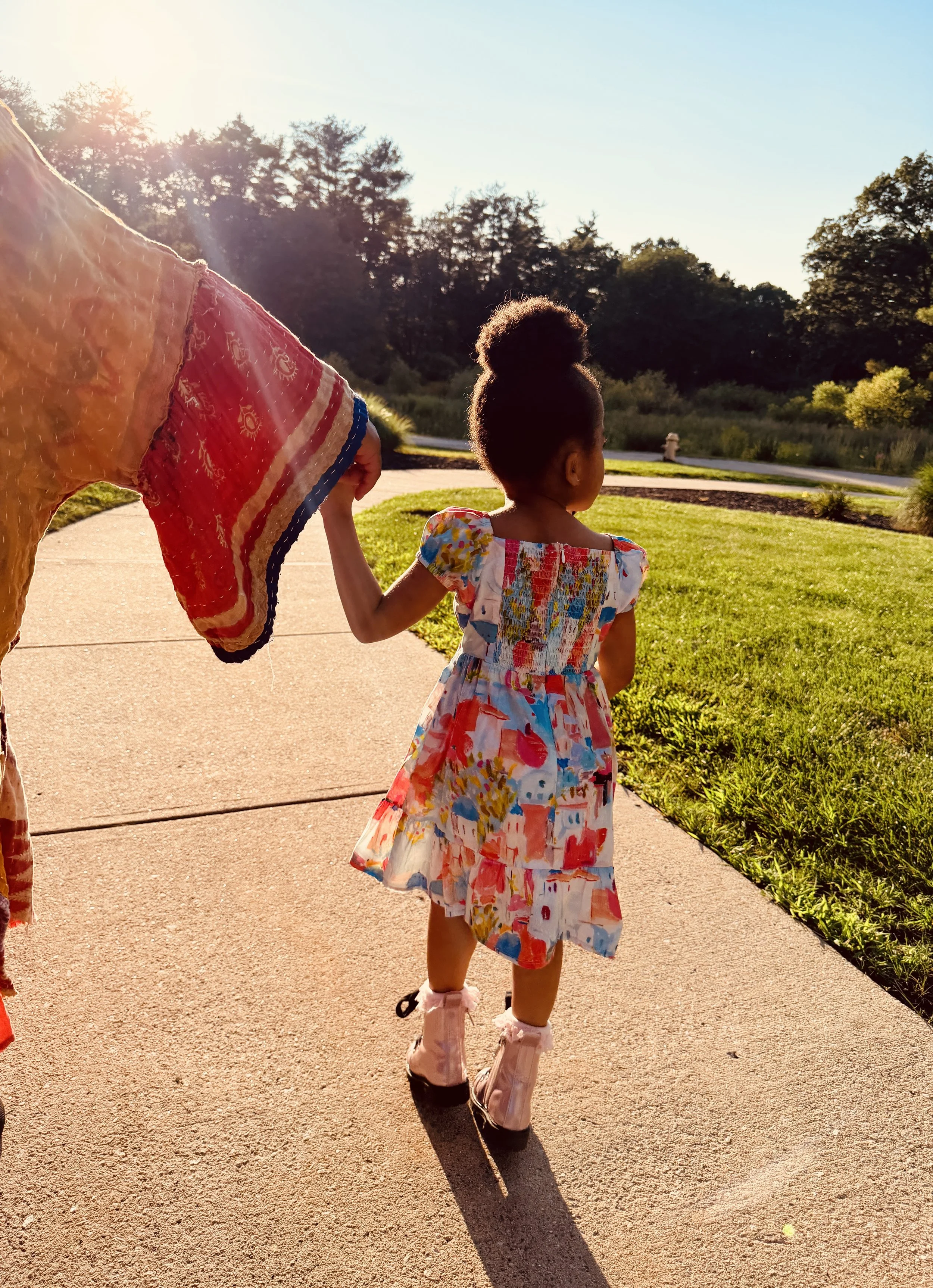 Young girl with her hair in a bun walking outdoors on a sunny day, holding an adult's hand, wearing a colorful floral dress and pink boots, with a grassy park and trees in the background.