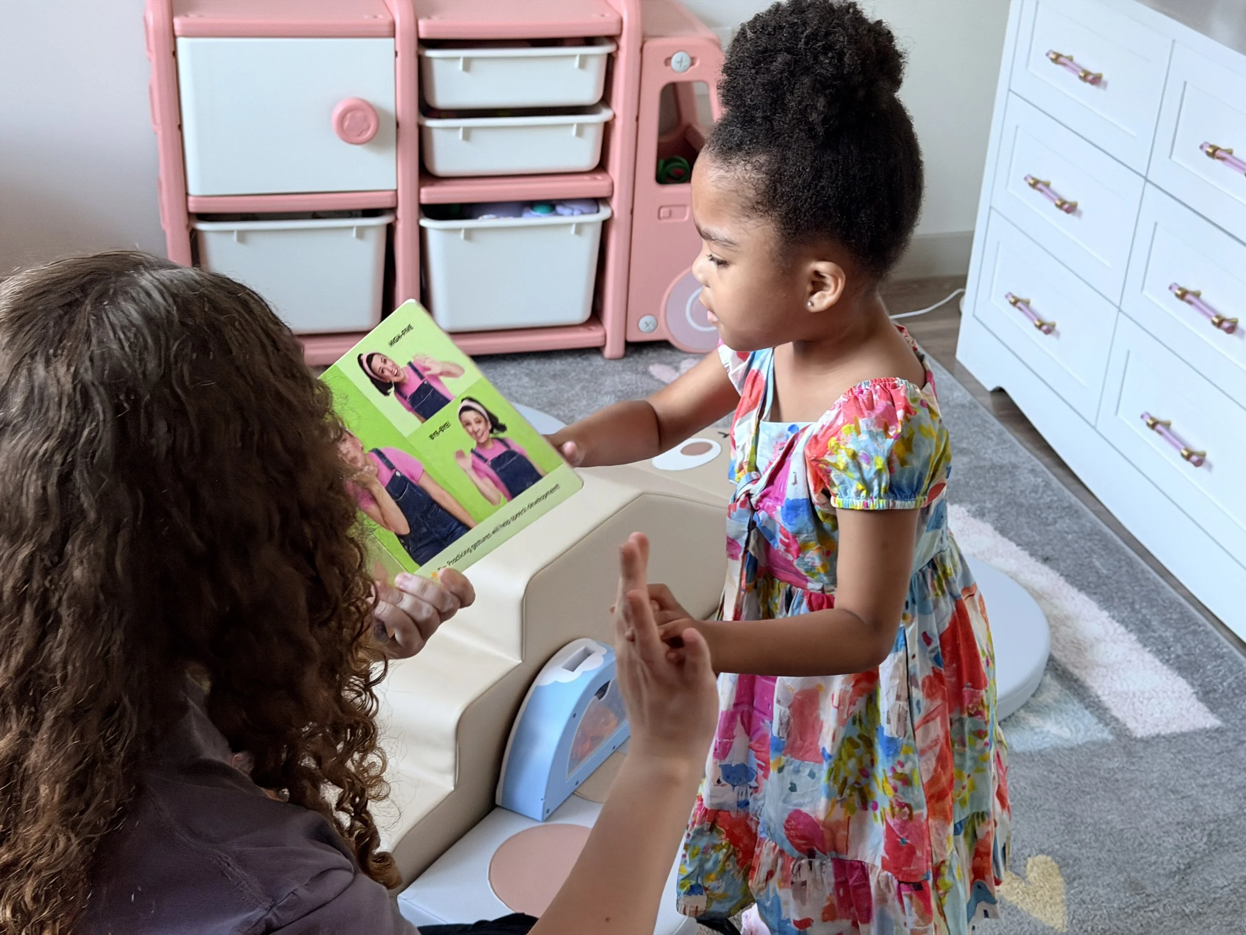 A young girl with curly hair in a colorful dress is looking at a woman with curly hair who is holding a brightly colored book with pictures of girls on the pages. They are in a room with white and pink furniture, including a pink toy kitchen and a white chest of drawers.