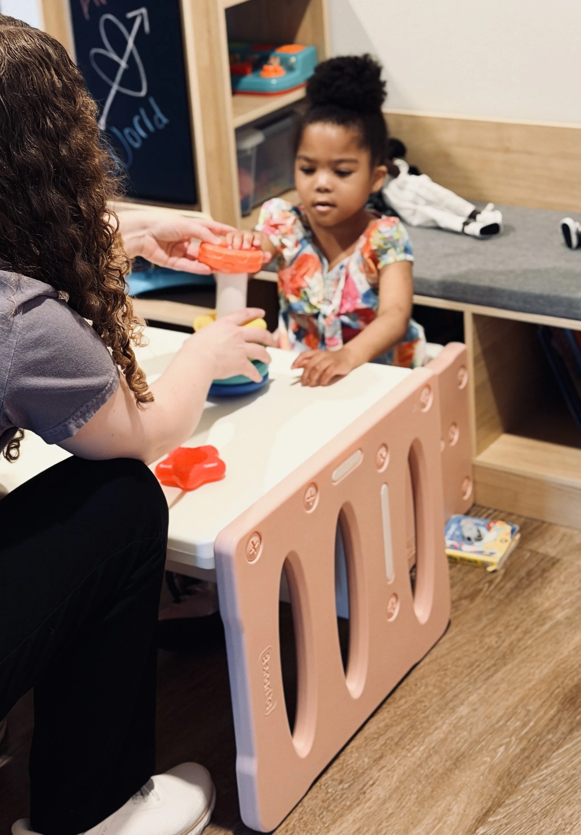 A young girl with curly hair wearing a colorful floral dress playfully interacts with an adult, who is handing her a toy, in a room with a pink play kitchen and some outside toys on the floor.