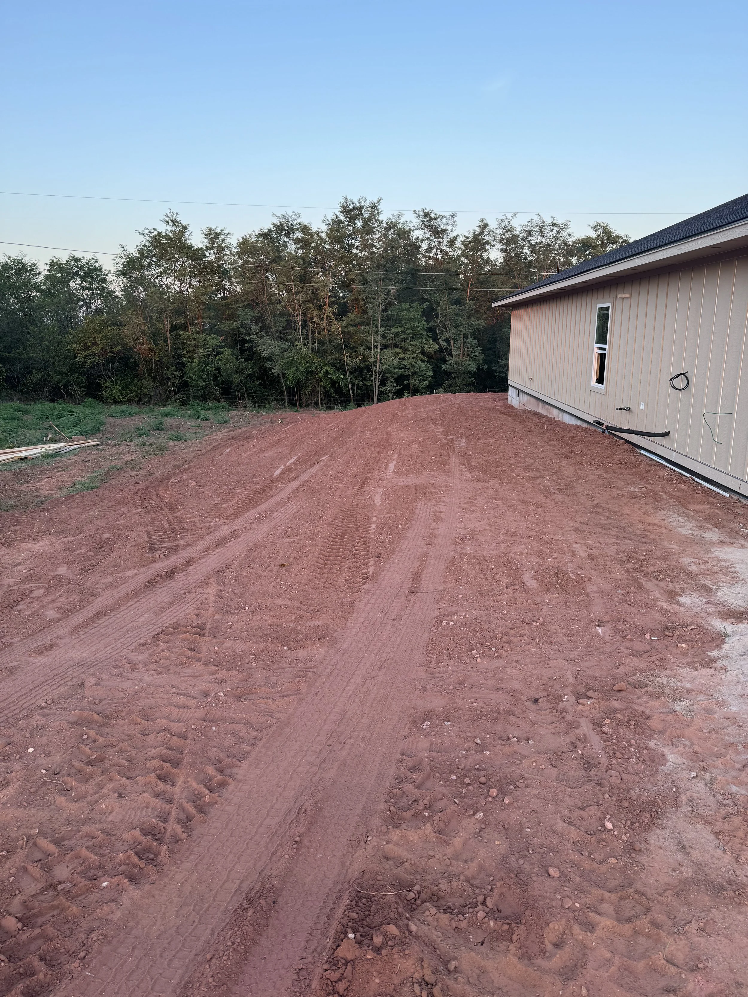 Dirt driveway beside a beige house with a window, with trees in the background under a clear sky.