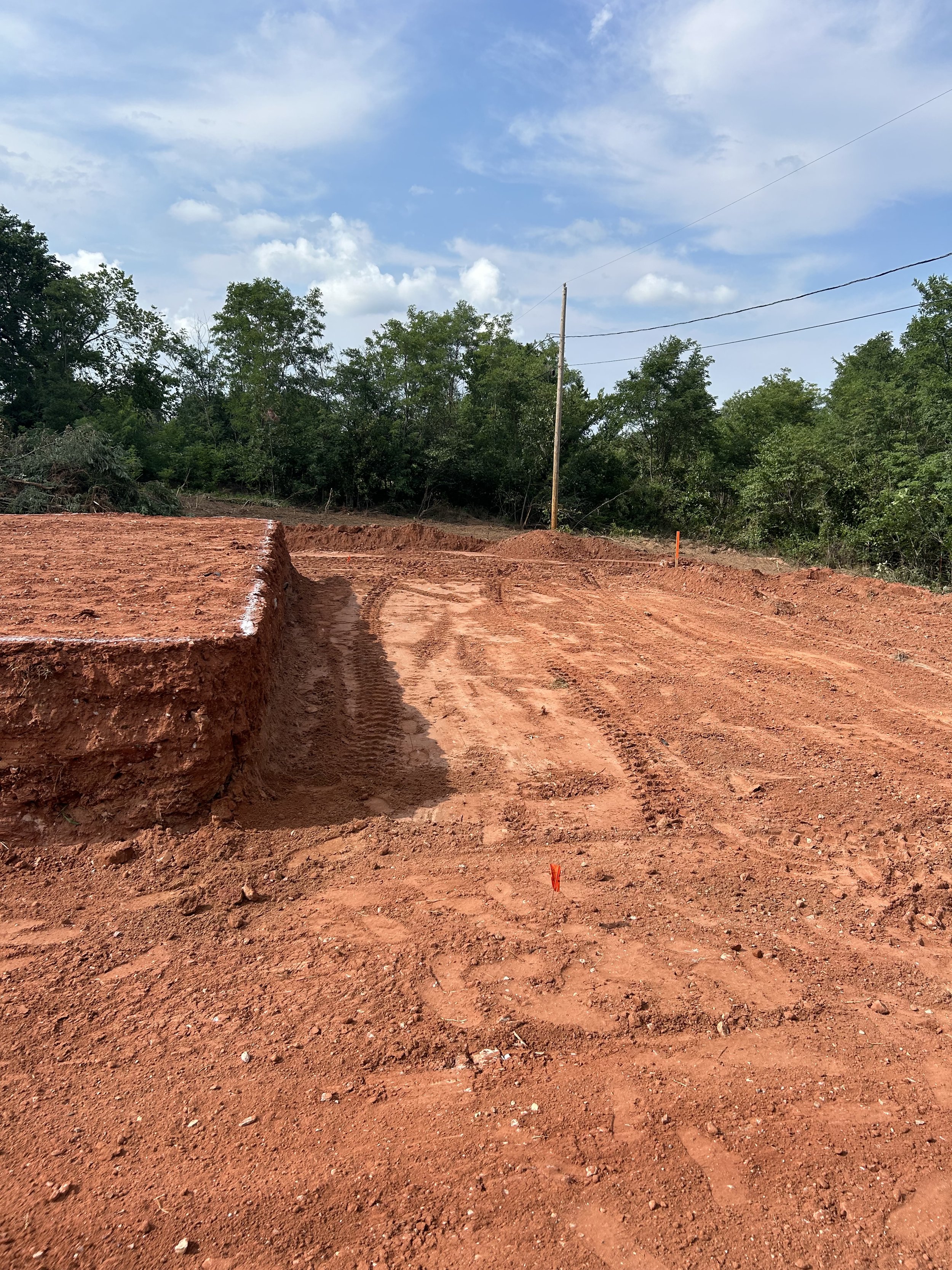 A construction site with red dirt, tire tracks, orange markers, green trees, utility pole, power lines, and a blue sky with clouds.