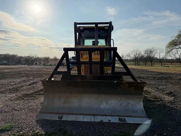 A large bulldozer with a metal blade in front, parked on a dirt field under a partly cloudy sky.