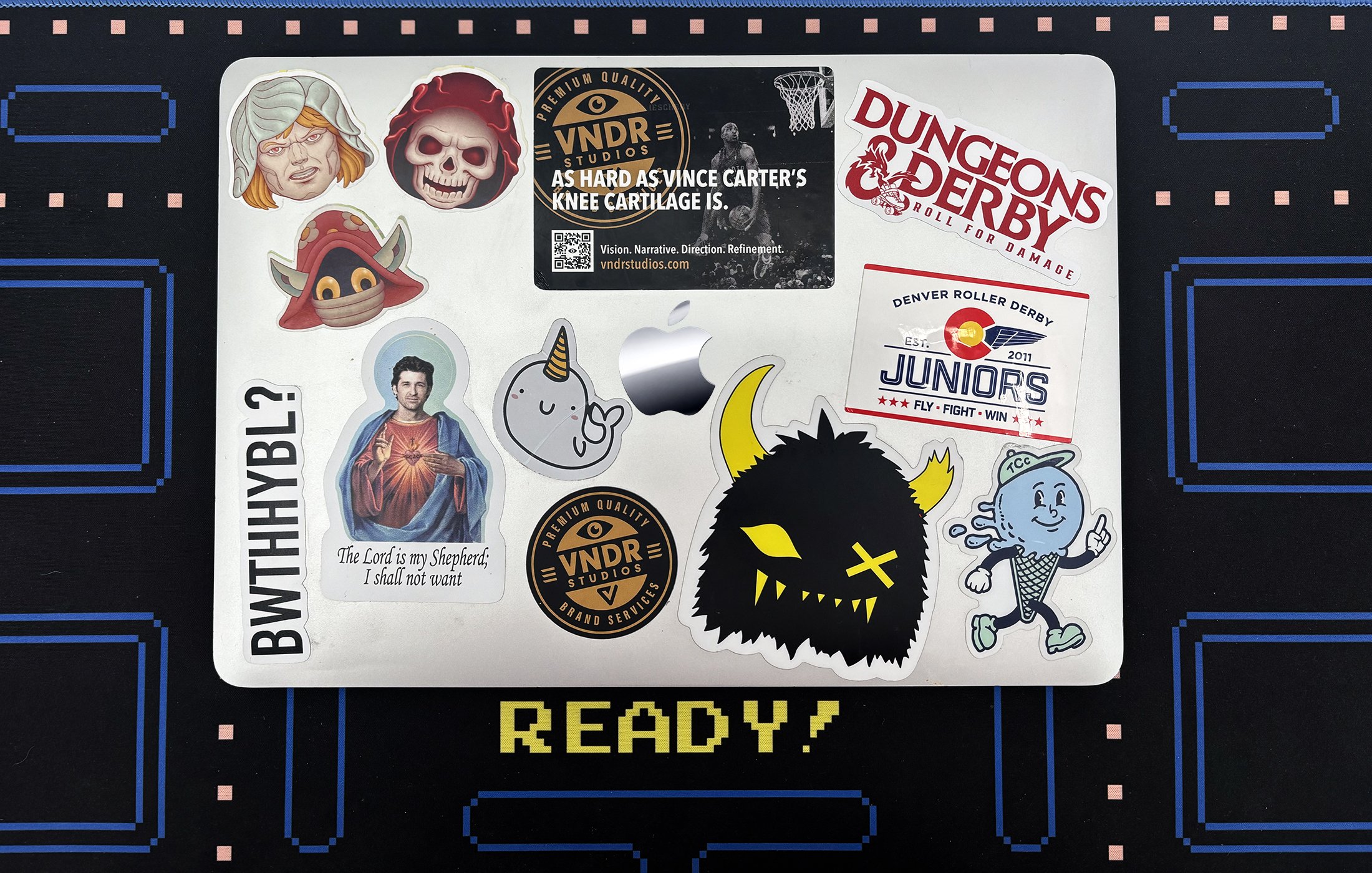 A laptop cover decorated with various stickers, including cartoon characters, gaming references, a religious quote, and event logos, over a background with a pixelated video game style design.