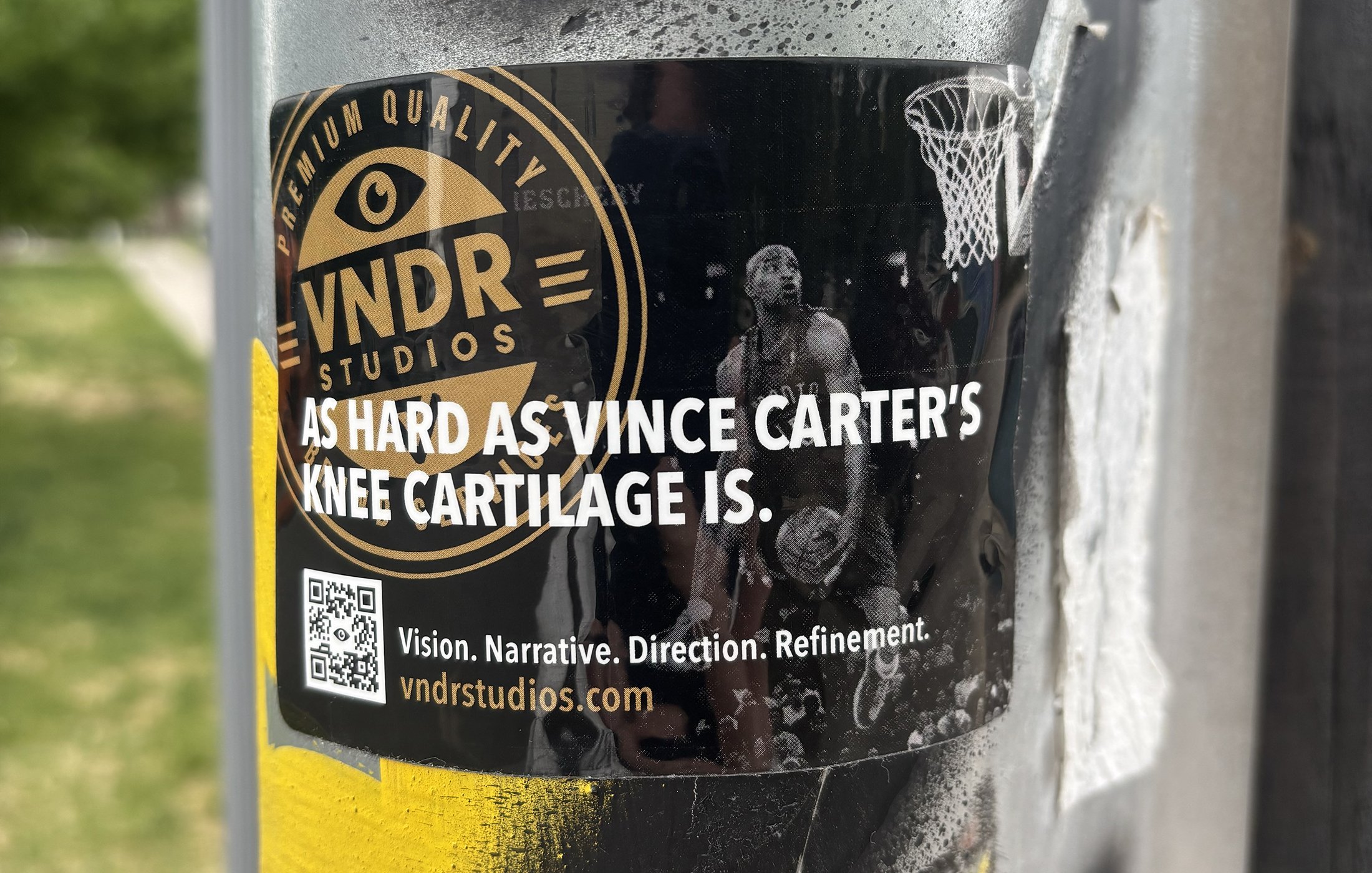 Close-up of a black sticker on a metal pole featuring the logo for VND Studio, with a basketball player image and the text 'As hard as Vince Carter's knee cartilage is.' The sticker also includes a QR code and website URL.