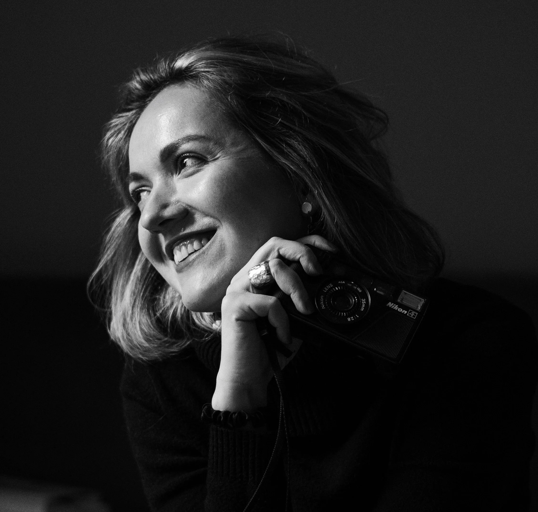 A black and white close-up portrait of a smiling woman with tousled hair holding a camera, wearing rings and a bracelet. Copyright Dasha Dare.