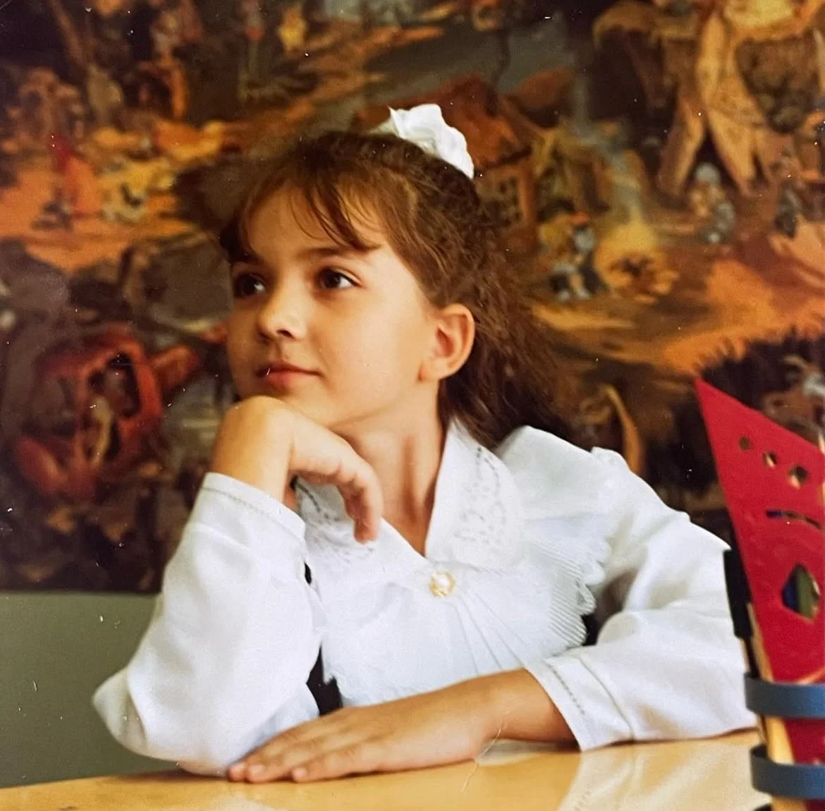 A young girl with long, curly brown hair and a white blouse with lace details, resting her chin on her hand, looking thoughtfully to her left. In the background, there is a detailed farm scene mural depicting barns, animals, and people. Copyright Das