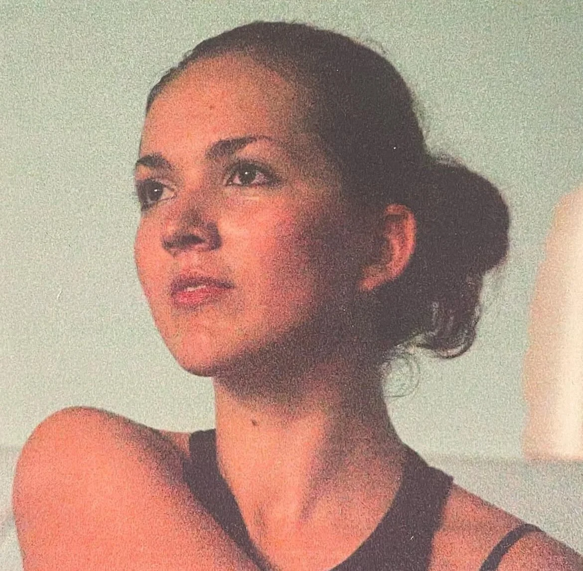 A woman with dark hair in a bun, wearing a sleeveless black top, looking to the side against a plain background. Copyright Dasha Dare.
