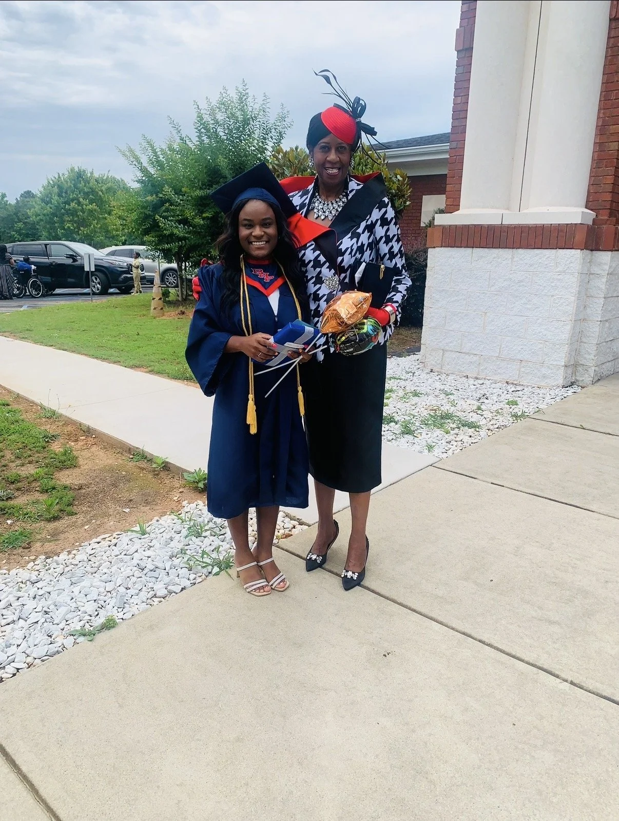 A young woman in a navy blue graduation gown and cap stands next to a woman dressed in stylish formal attire, both smiling and holding certificates and balloons outside a building with a lawn and parking lot in the background.