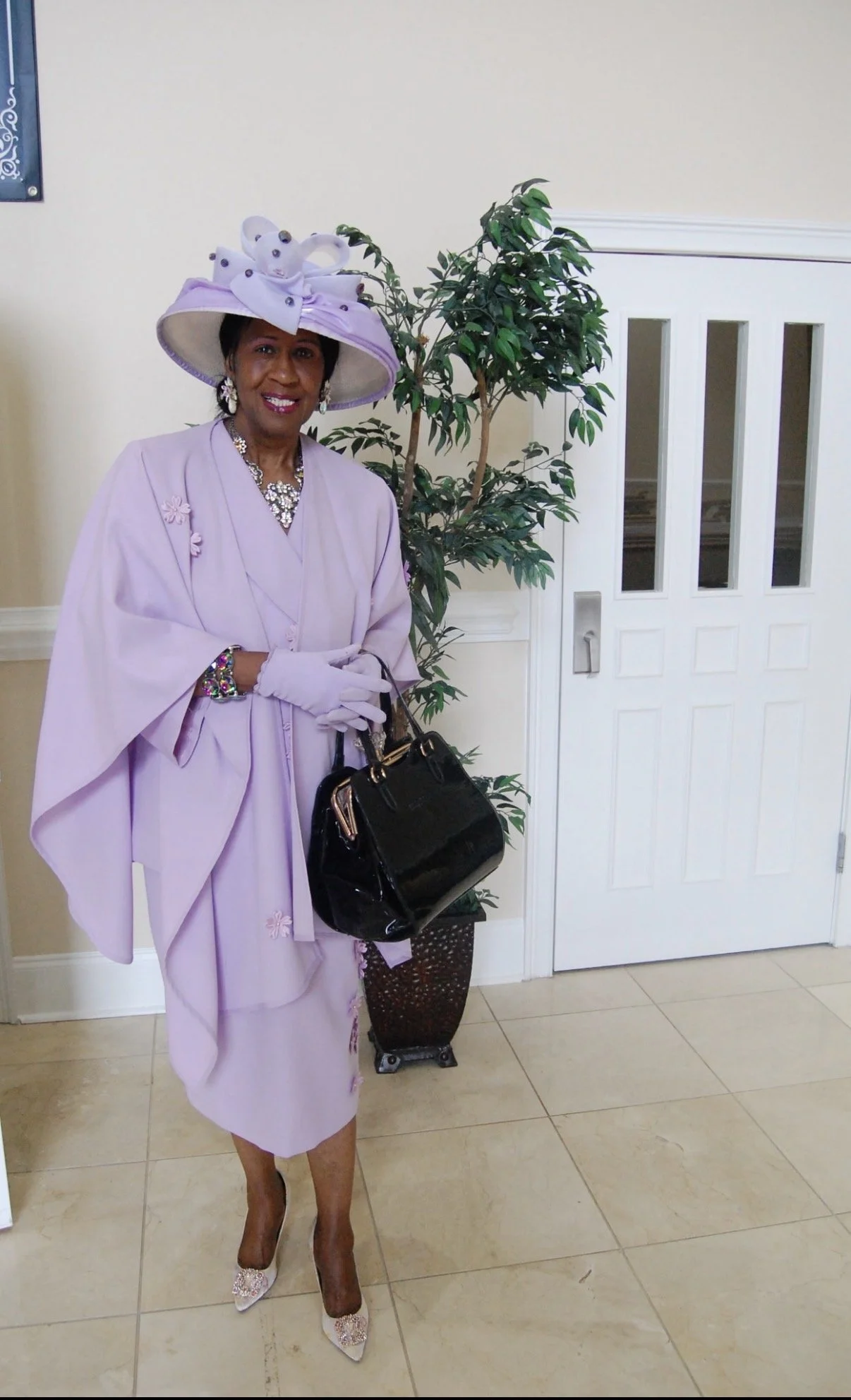 A woman dressed in a lavender outfit with a large matching hat, standing indoors next to a potted plant and a white door.